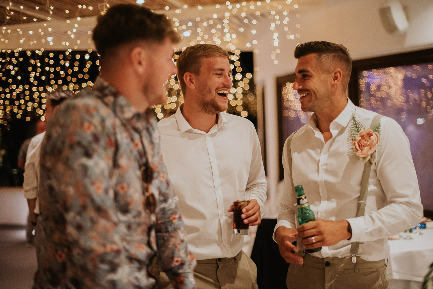 Wedding photographer Santorini: groom and his friends chatting and laughing under the fairy lights by Ben and Vesna.