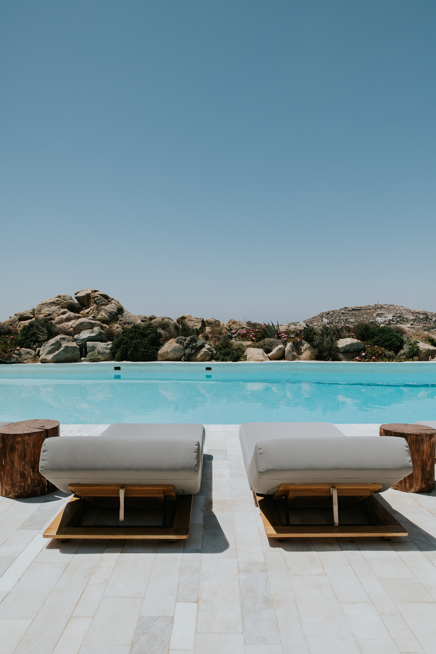 Mykonos wedding photographer: sun beds await at the pool with its turquoise water surrounded by Mykonian rocks and wild herbs.