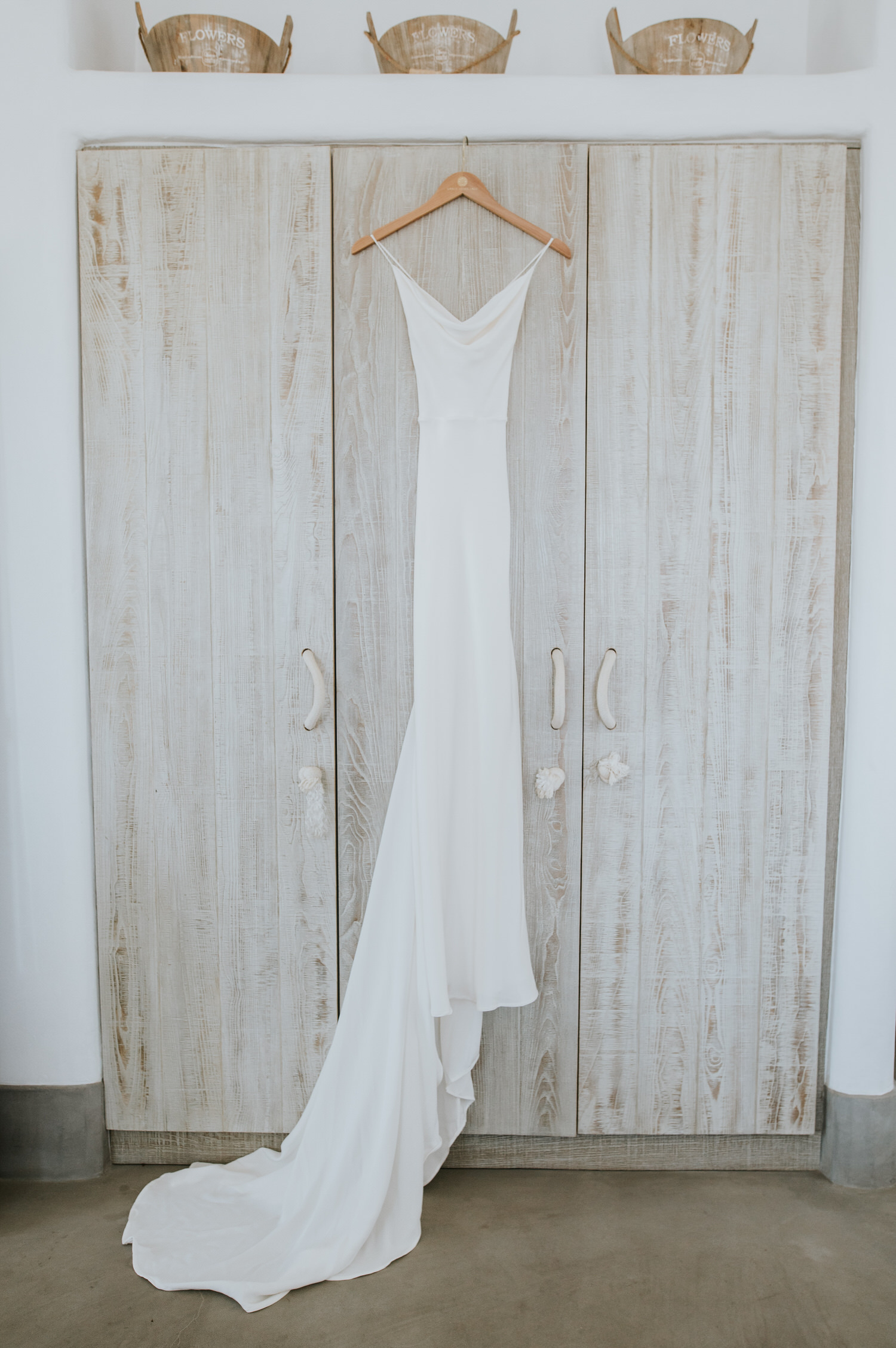 Mykonos wedding photographer: gorgeous floaty Grace loves lace wedding dress hanging on the beautiful wooden wardrobe door with three wooden baskets on top.