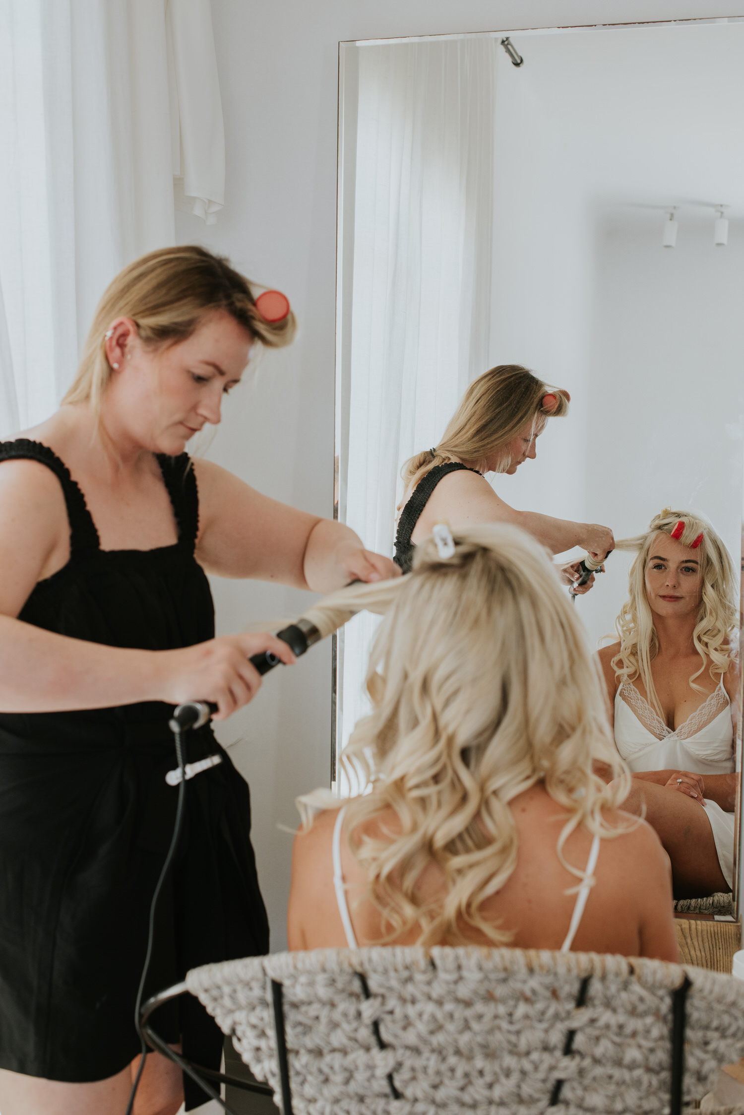 Mykonos wedding photographer: bride sat on a chair looking at herself in the mirror as her bestie does her hair for Mykonos wedding.