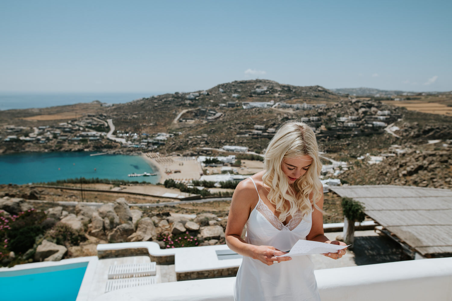 Mykonos wedding photographer: bride leaning on the wall on the terrace with the beach in the background reading a note her husband to be sent.