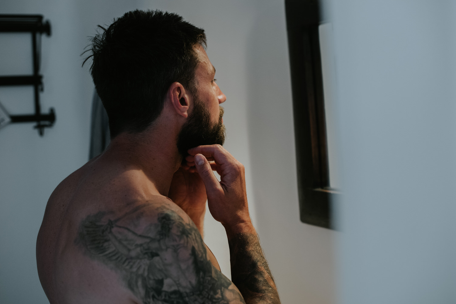 Mykonos wedding photographer: side portrait of shirtless groom looking in the mirror fixing his beard.