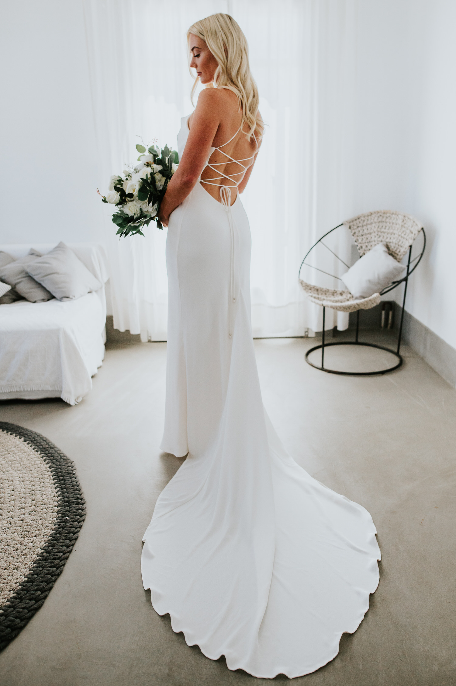 Mykonos wedding photographer: gorgeous bride holding bouquet showing her back of the wedding dress in the room.