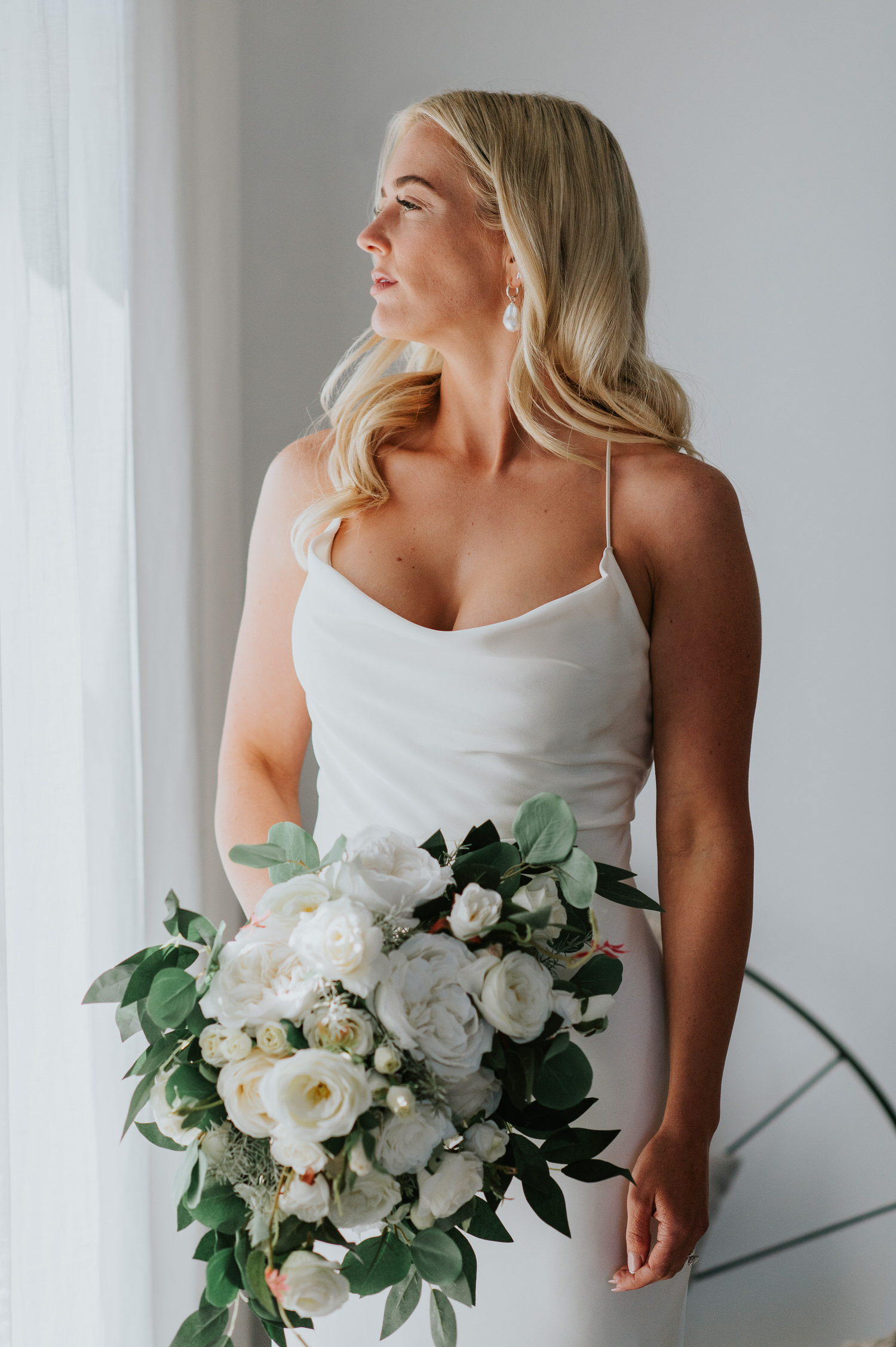 Mykonos wedding photographer: stunning bride looking out through the window holding bouquet in beautiful light.