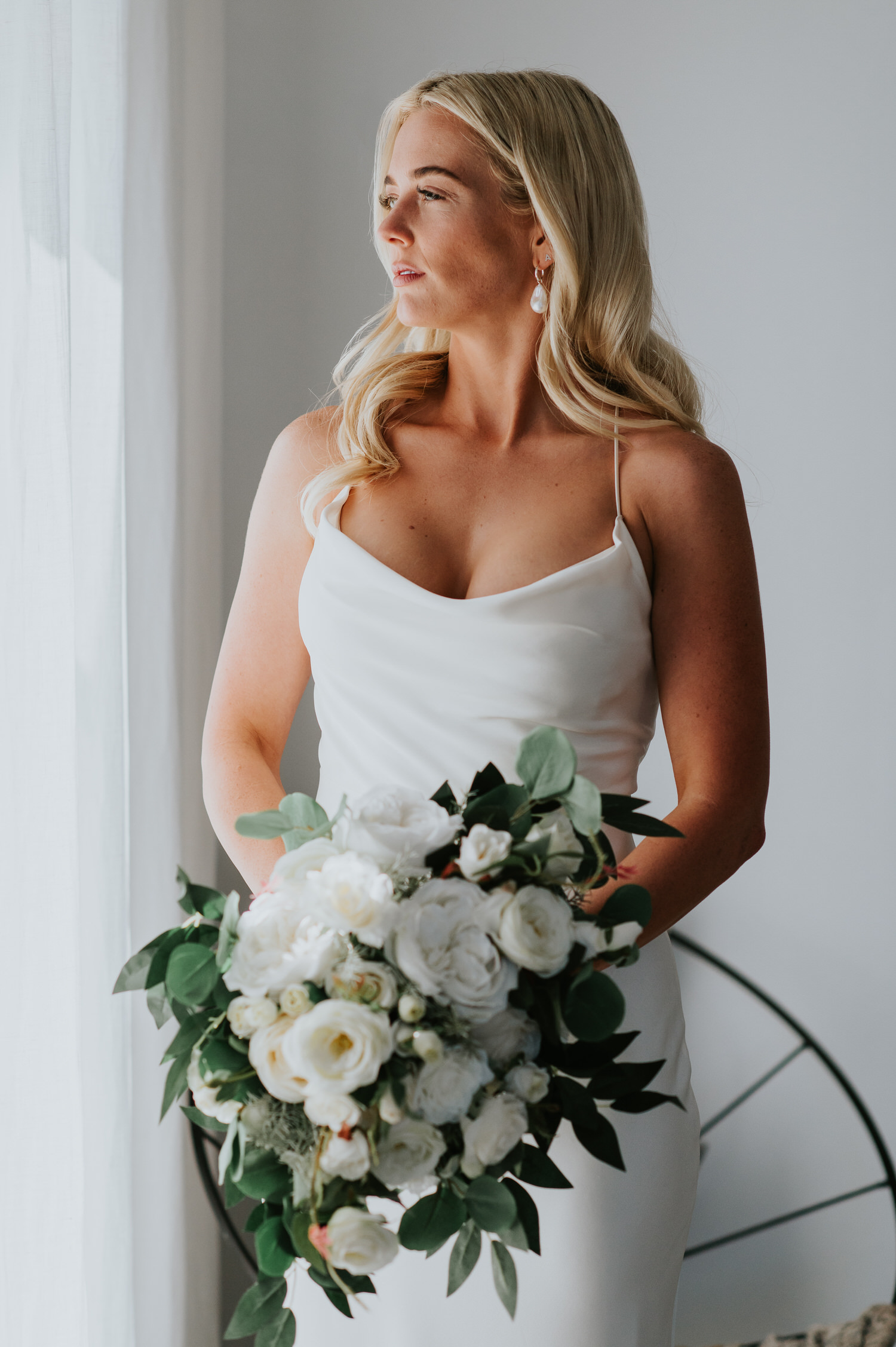 Mykonos wedding photographer: stunning bride looking out through the window holding her bouquet in beautiful light.