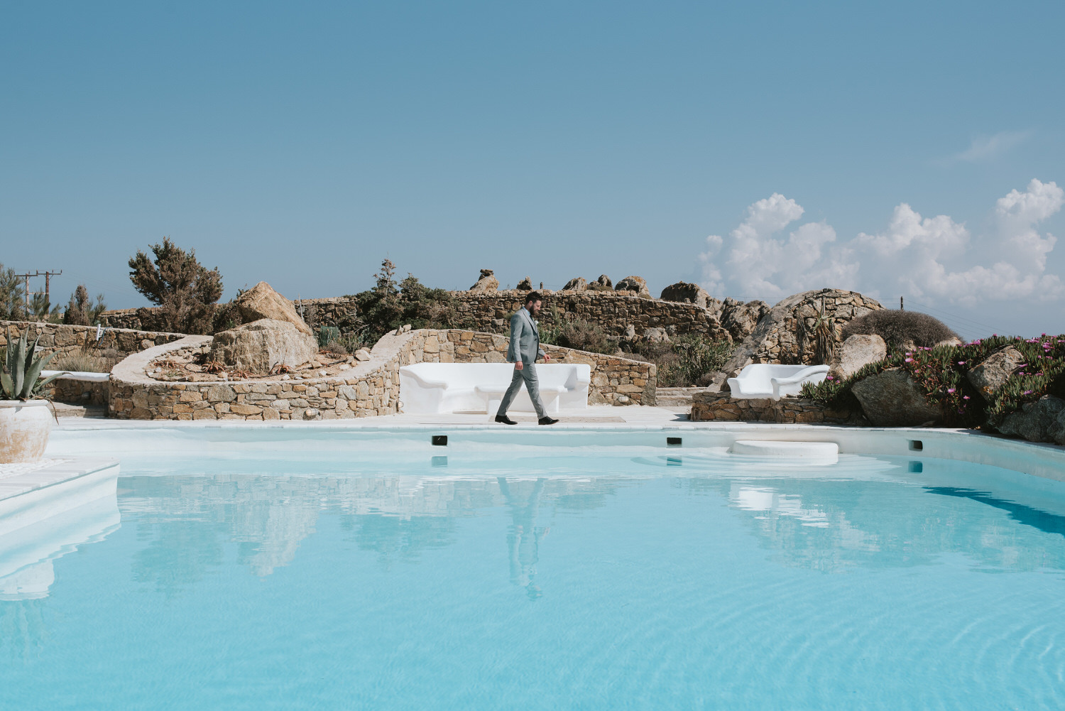 Mykonos wedding photographer: groom walking on the other side of the pool with water reflections before the first look and the rocks in the background.
