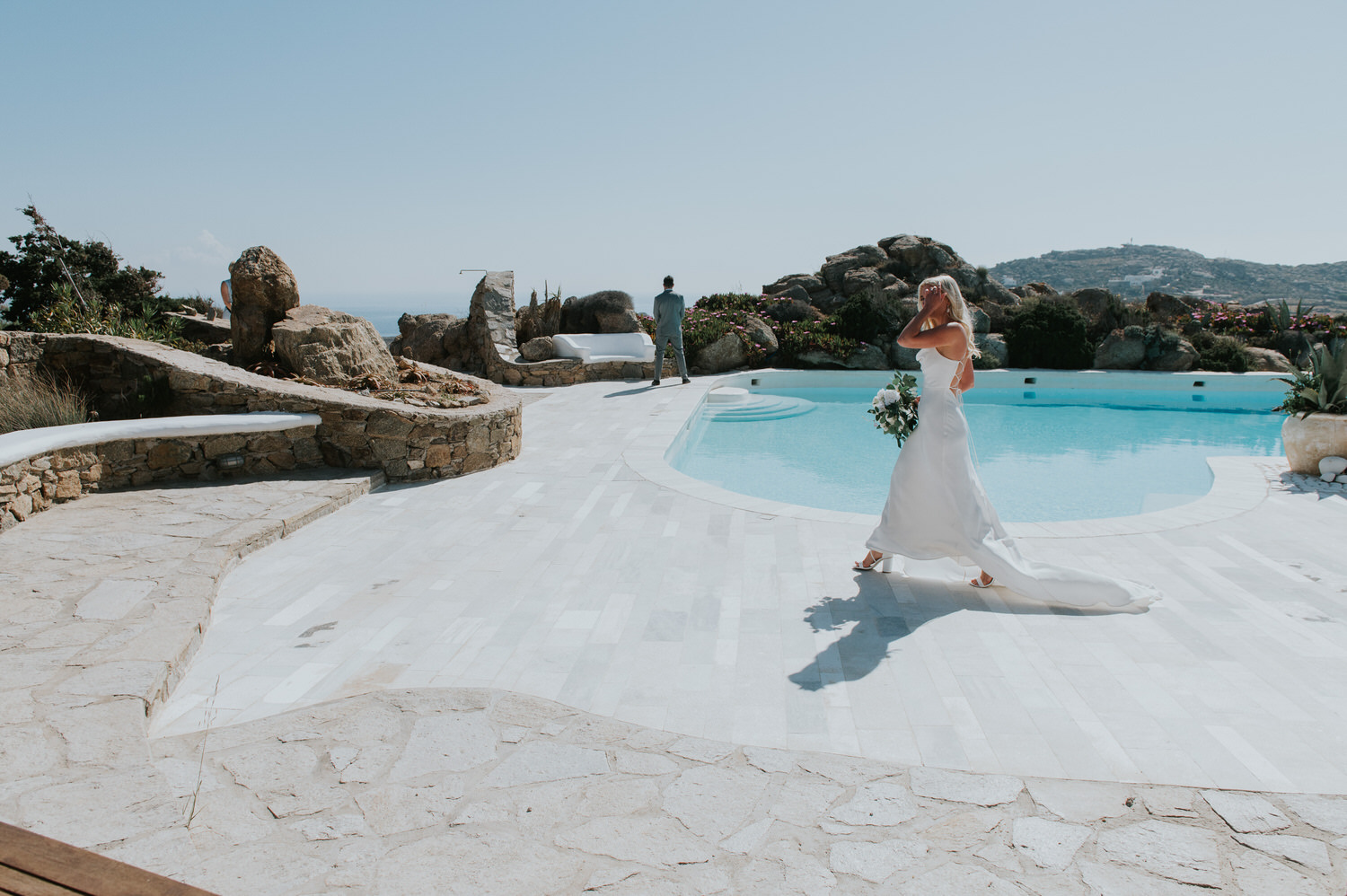 Mykonos wedding photographer: bride walking to her groom next to the pool with his back to her for their first look.