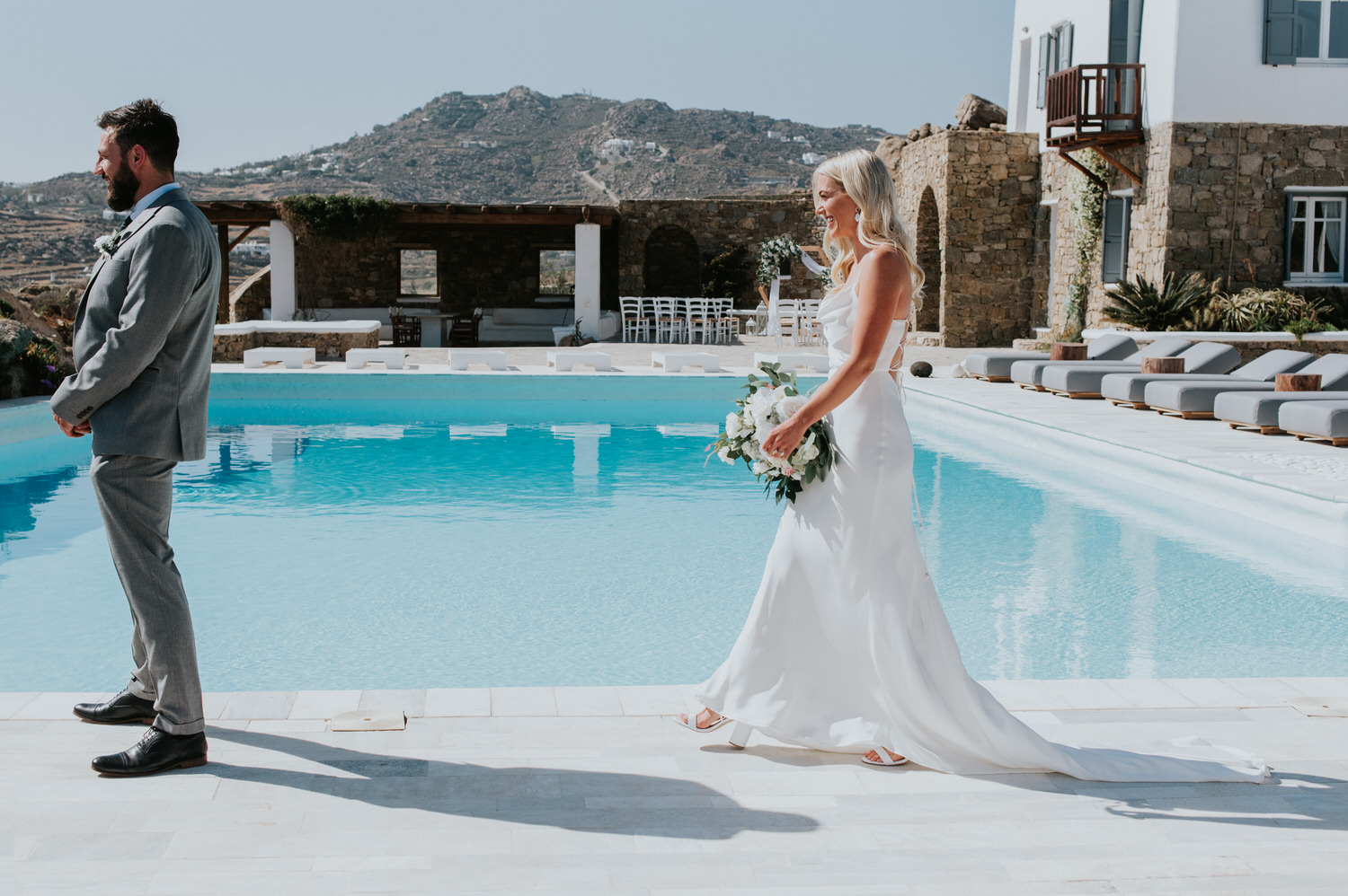Mykonos wedding photographer: close up of bride smiling walking to her groom next to the pool with his back to her for their first look.