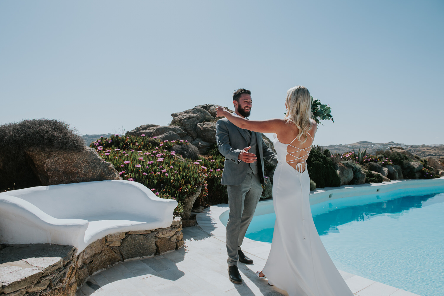 Mykonos wedding photographer: bride and groom going for a hug during first look next to the pool and the rocks in the background.