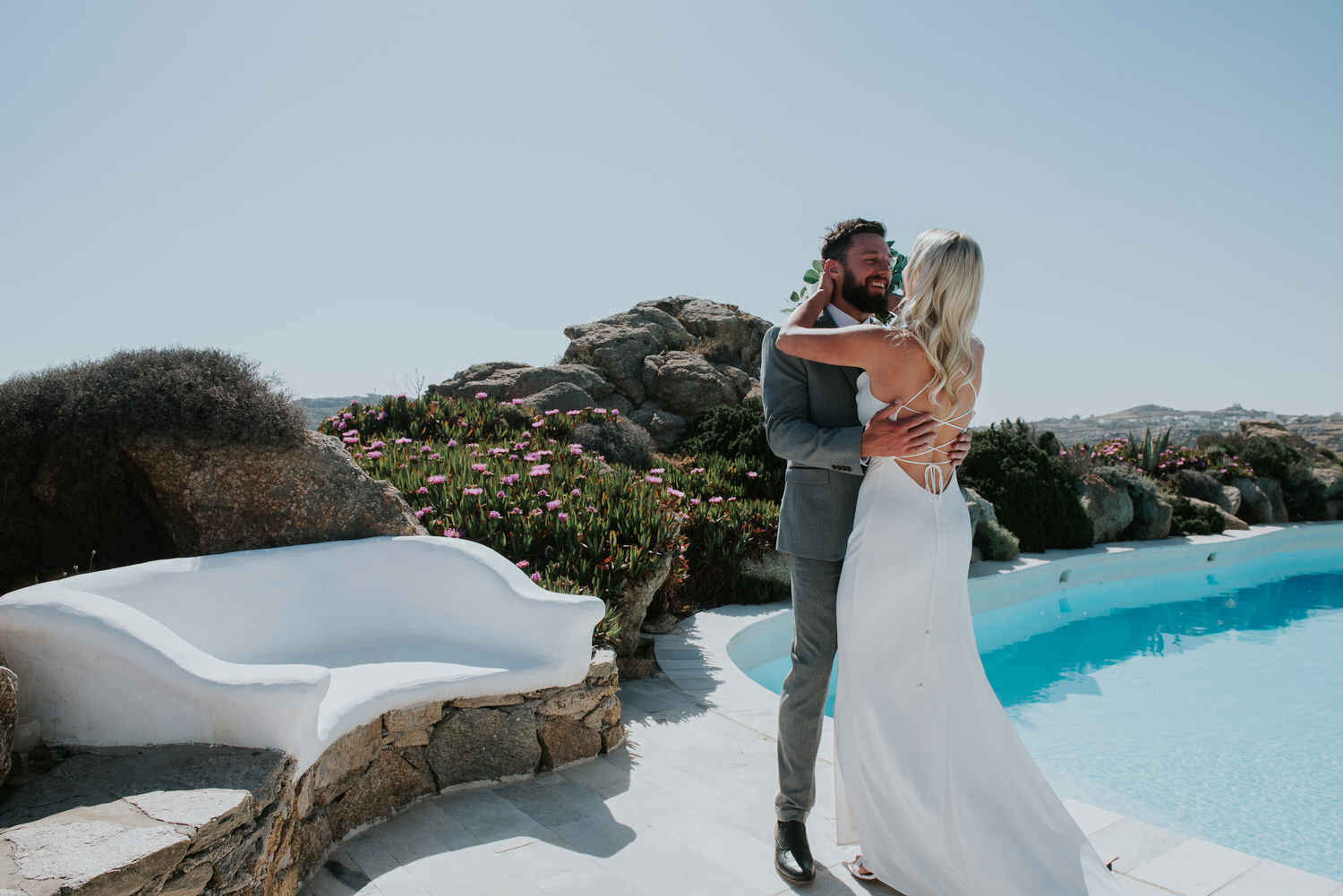 Mykonos wedding photographer: bride reaches out to her groom next to the pool and the rocks in the background.
