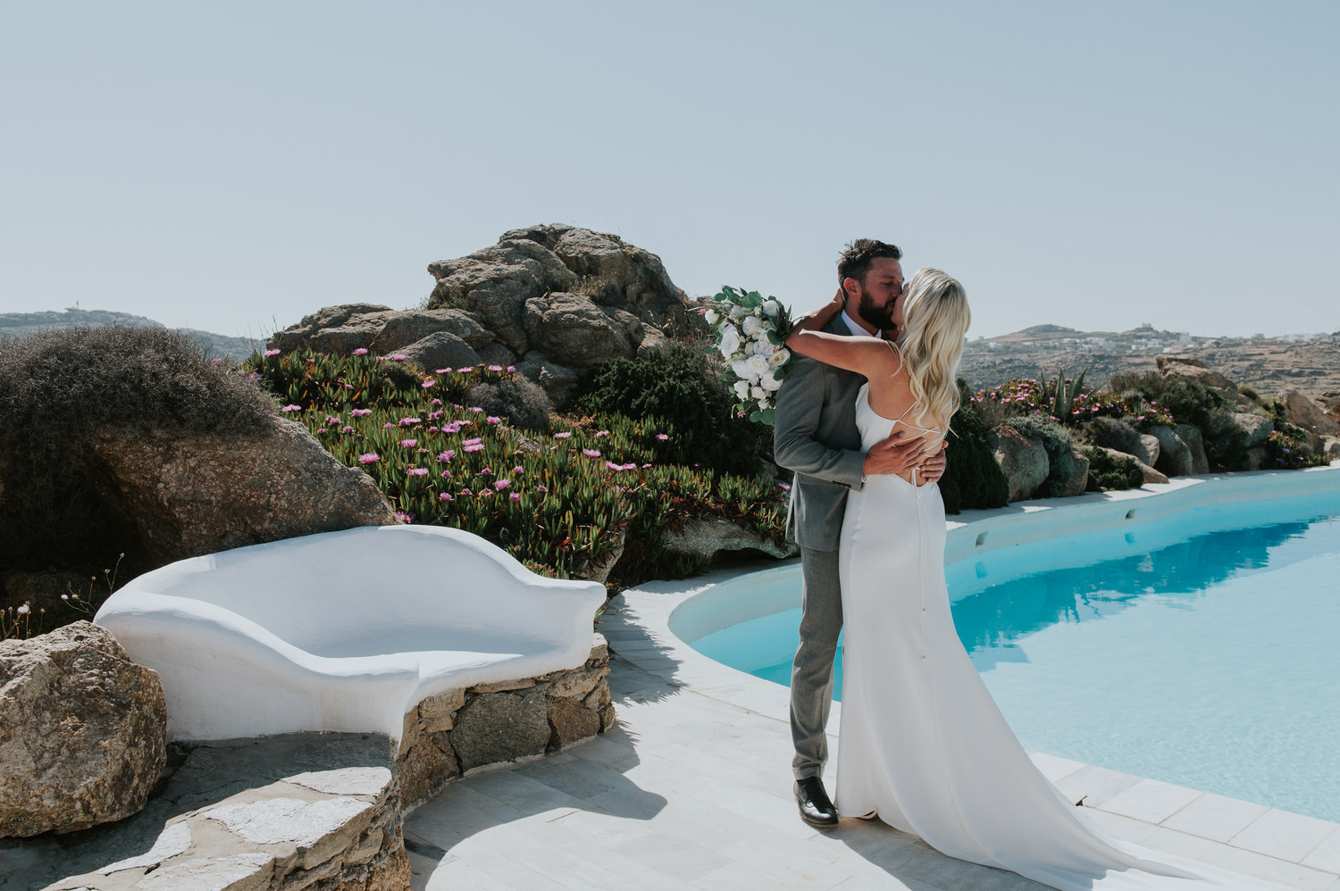 Mykonos wedding photographer: bride and groom embraced in a kiss next to the pool and the rocks in the background.