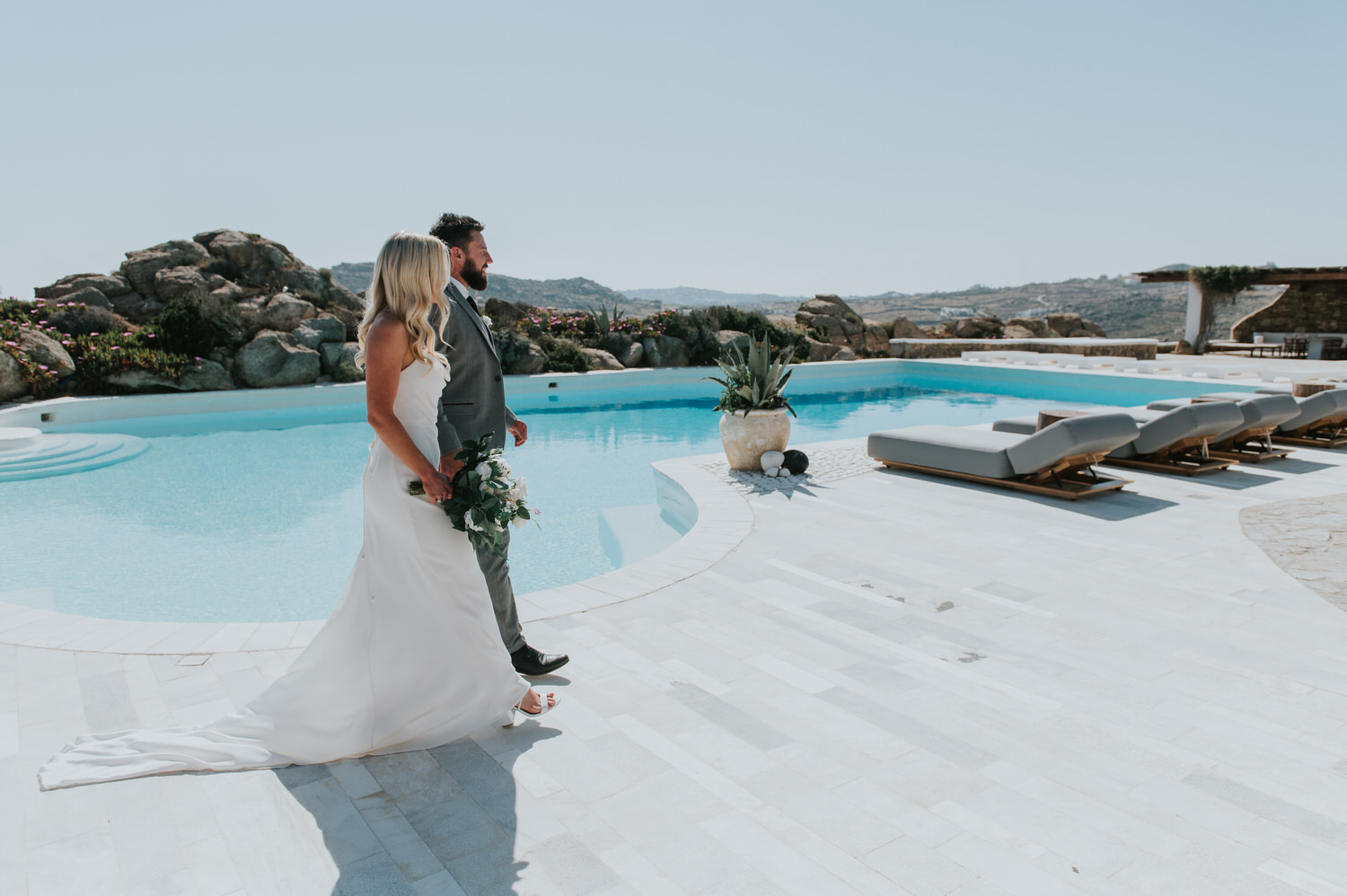 Mykonos wedding photographer: bride and groom walk on the terrace pass the swimming pool with the rocks in the background.