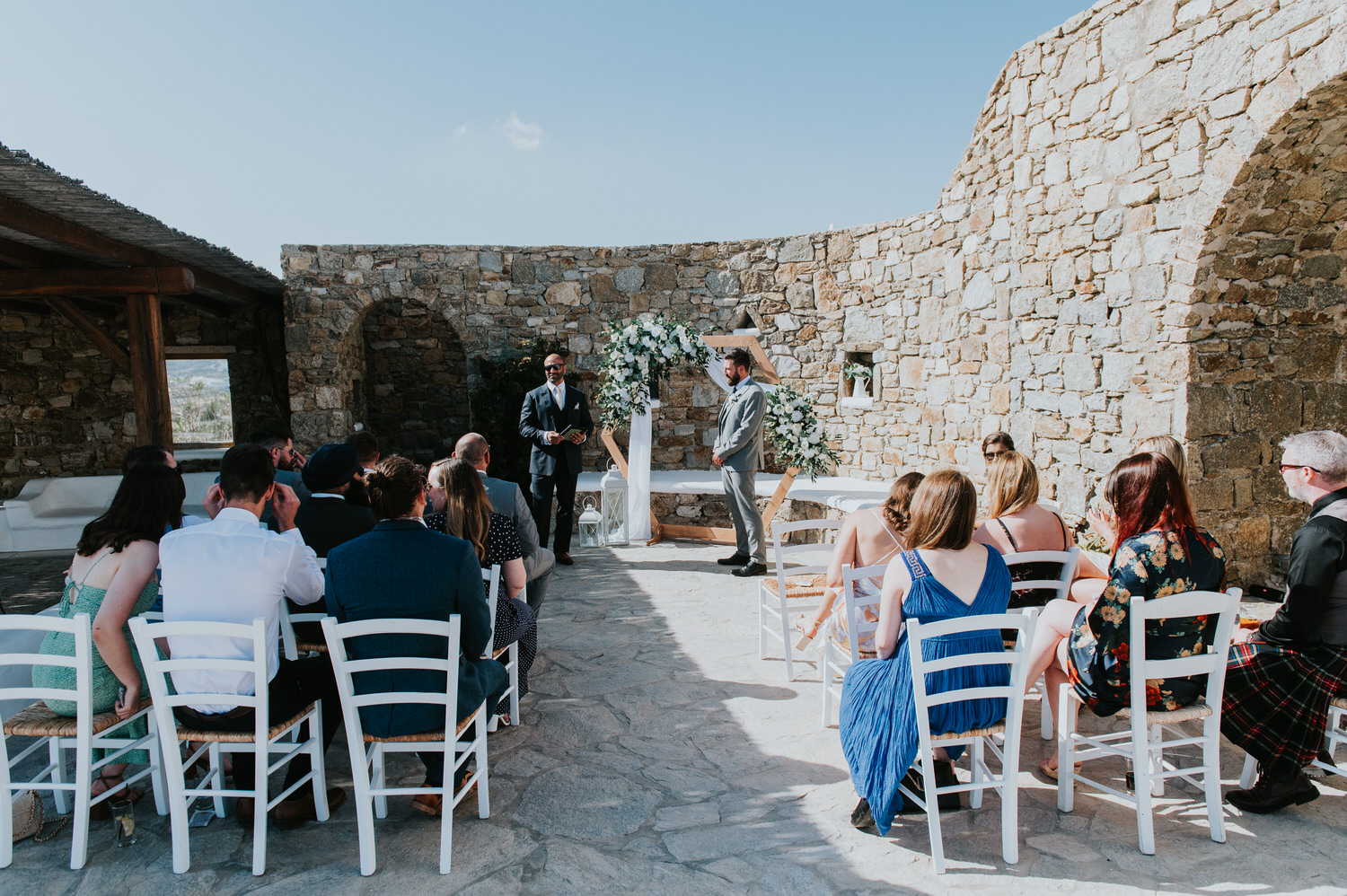 Mykonos wedding photographer: panoramic photo of the groom and the celebrant in front of the floral arch with guests in the foreground.