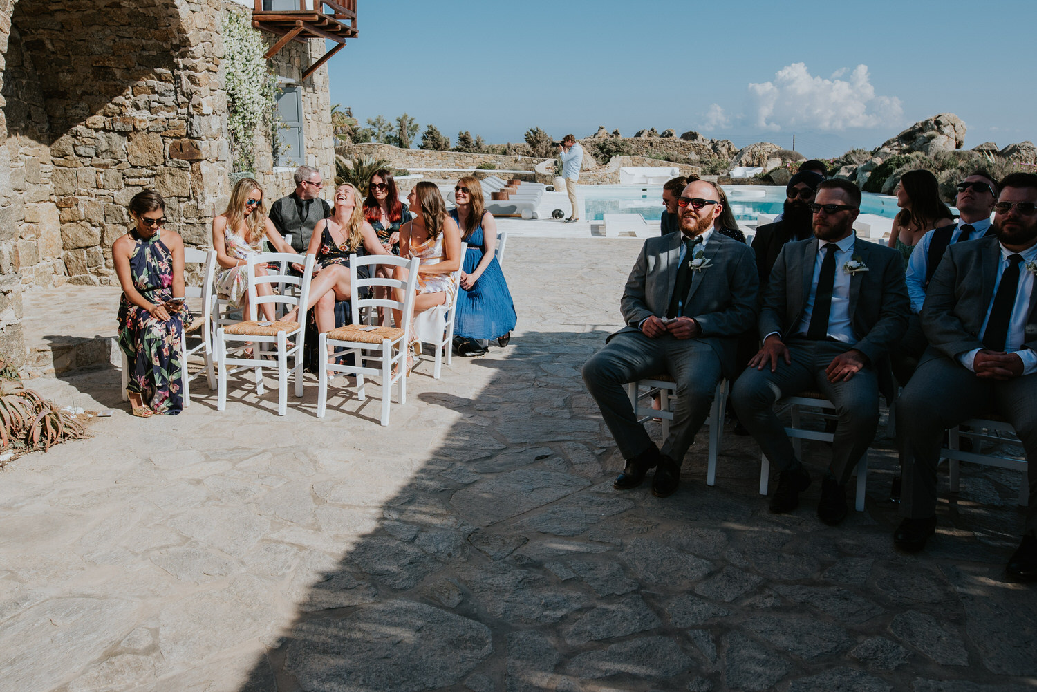 Guests sat at the ceremony terrace laughing and chatting waiting for the Mykonos wedding to start with Mykonos wedding photographer in the background.