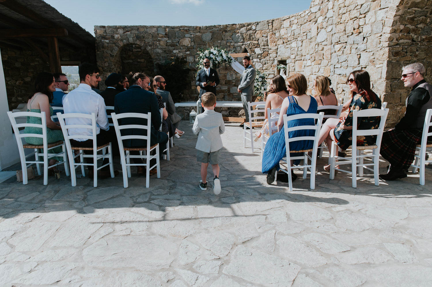 Mykonos wedding photographer: panoramic photo of the little ring bearer walking up the aisle as the guests look at him smiling.