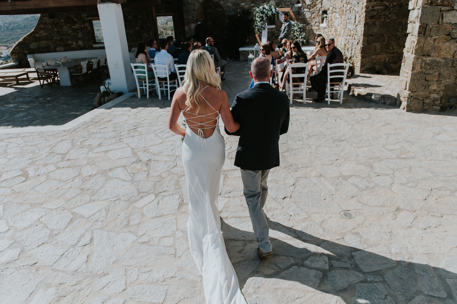 Mykonos wedding photographer: bride walking down the aisle in her beautiful dress with guests looking at her.