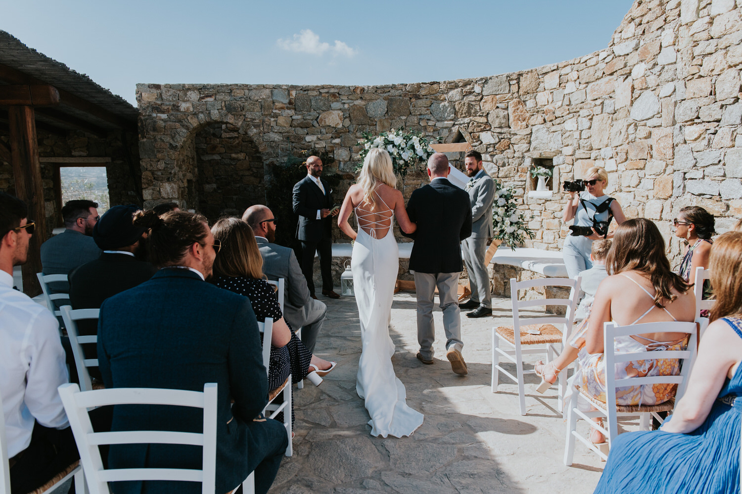 Mykonos wedding photographer: bride walking down the aisle as her beautiful dress' train trails behind her and the groom looks at her.