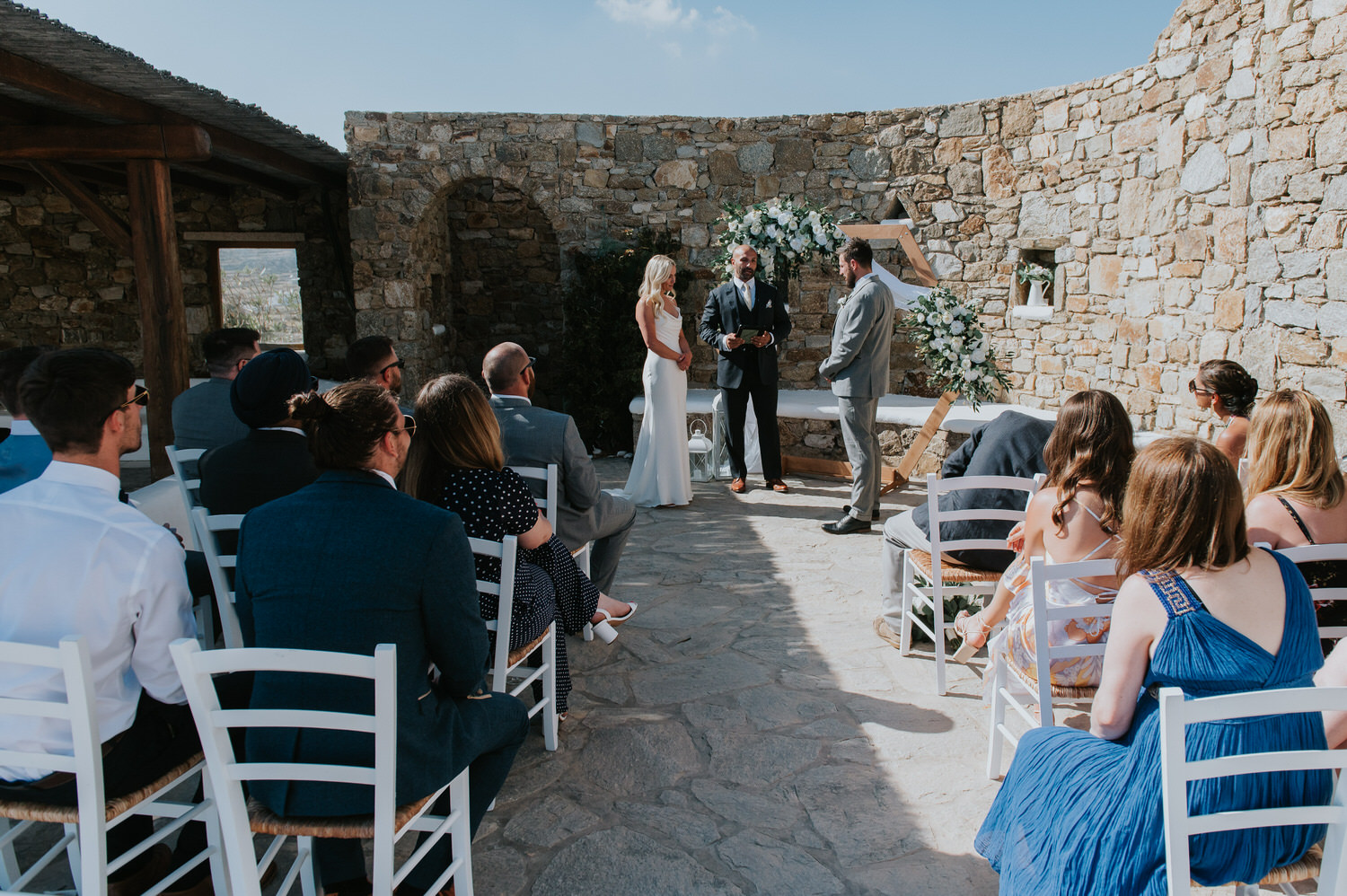 Mykonos wedding photographer: bride and groom stand in front of the the celebrant and the floral arch with guests sat in the foreground.
