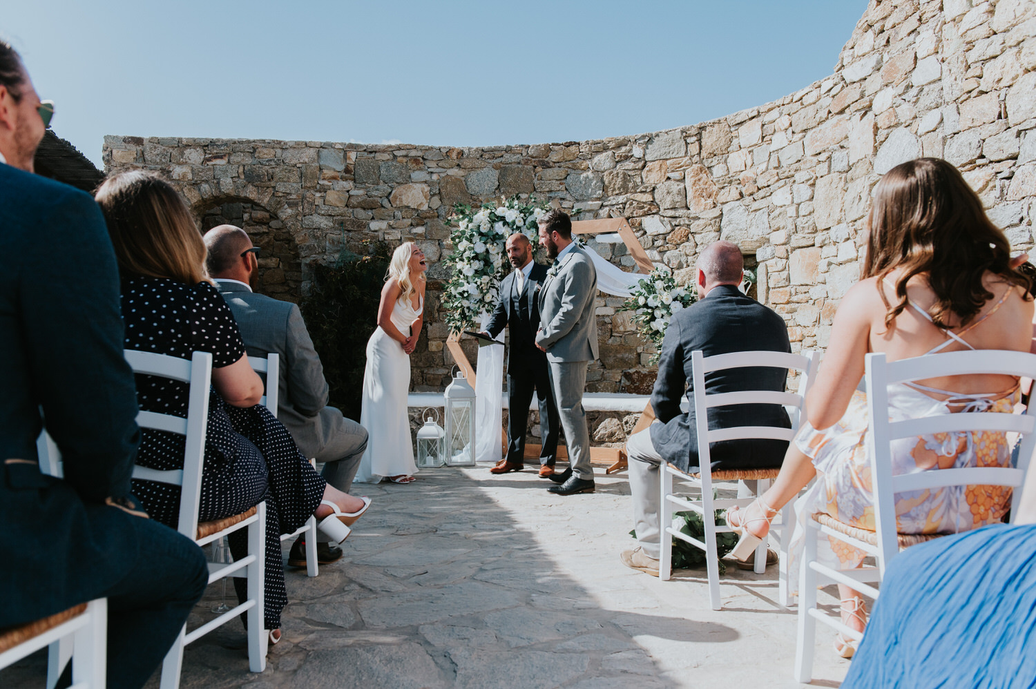 Mykonos wedding photographer: bride laughs as the celebrant and groom stand in front of the floral arch.