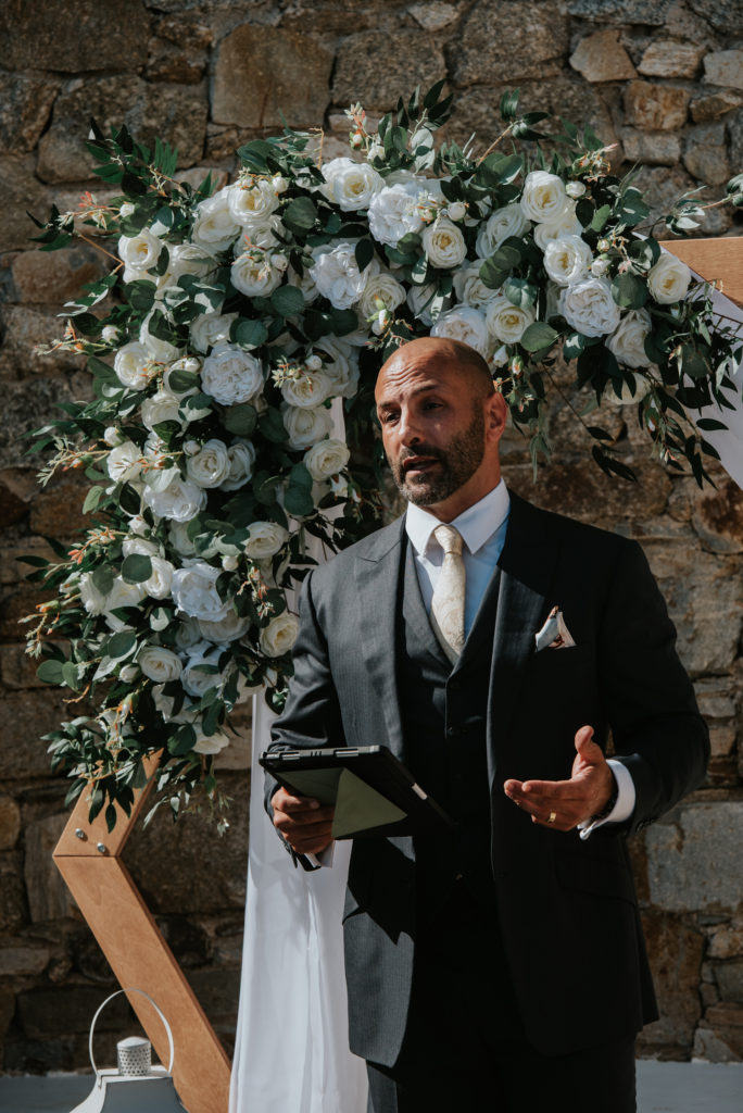 Mykonos wedding photographer: wedding celebrant looking handsome in front of the floral arch as the wedding ceremony starts.