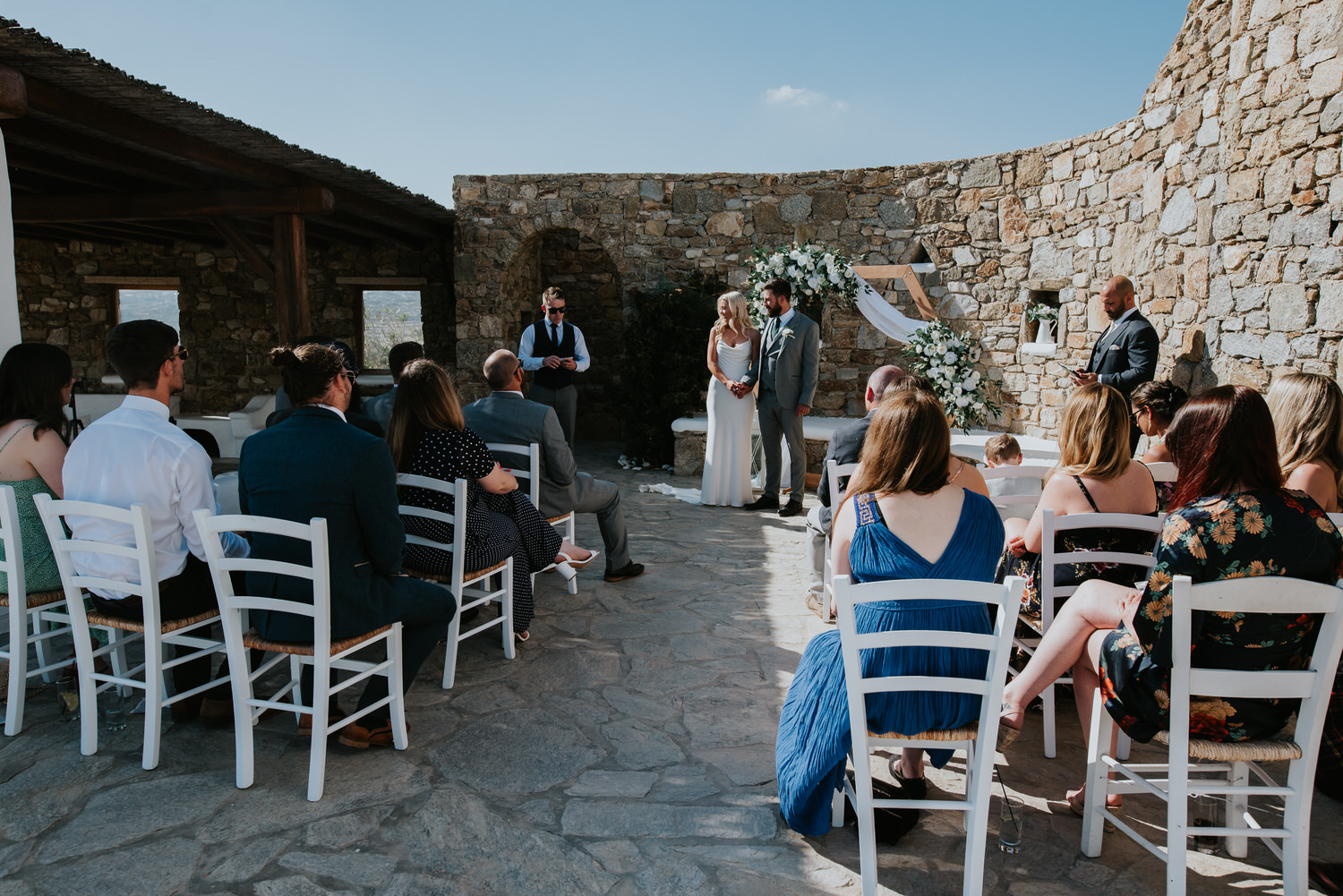 Mykonos wedding photographer: panoramic photo of bride and groom holding hands during the ceremony as they and the guests listen to a friend reading.