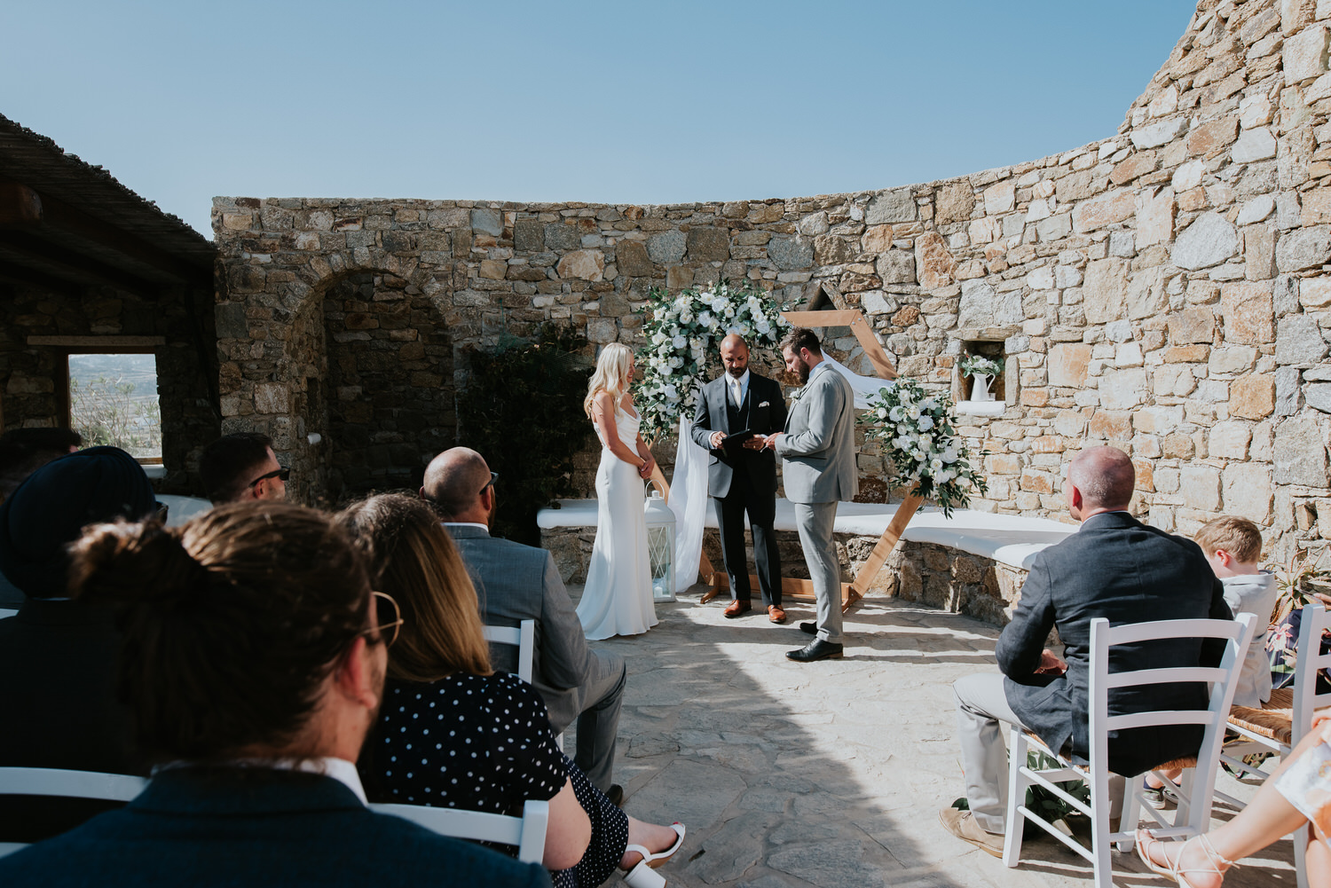 Mykonos wedding photographer: panoramic photo of bride and groom in front of the floral arch and the celebrant with guests sat in the foreground.