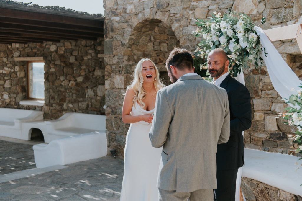 Mykonos wedding photographer: bride laughs as her groom's reads his vows in front of the floral arch and the celebrant.