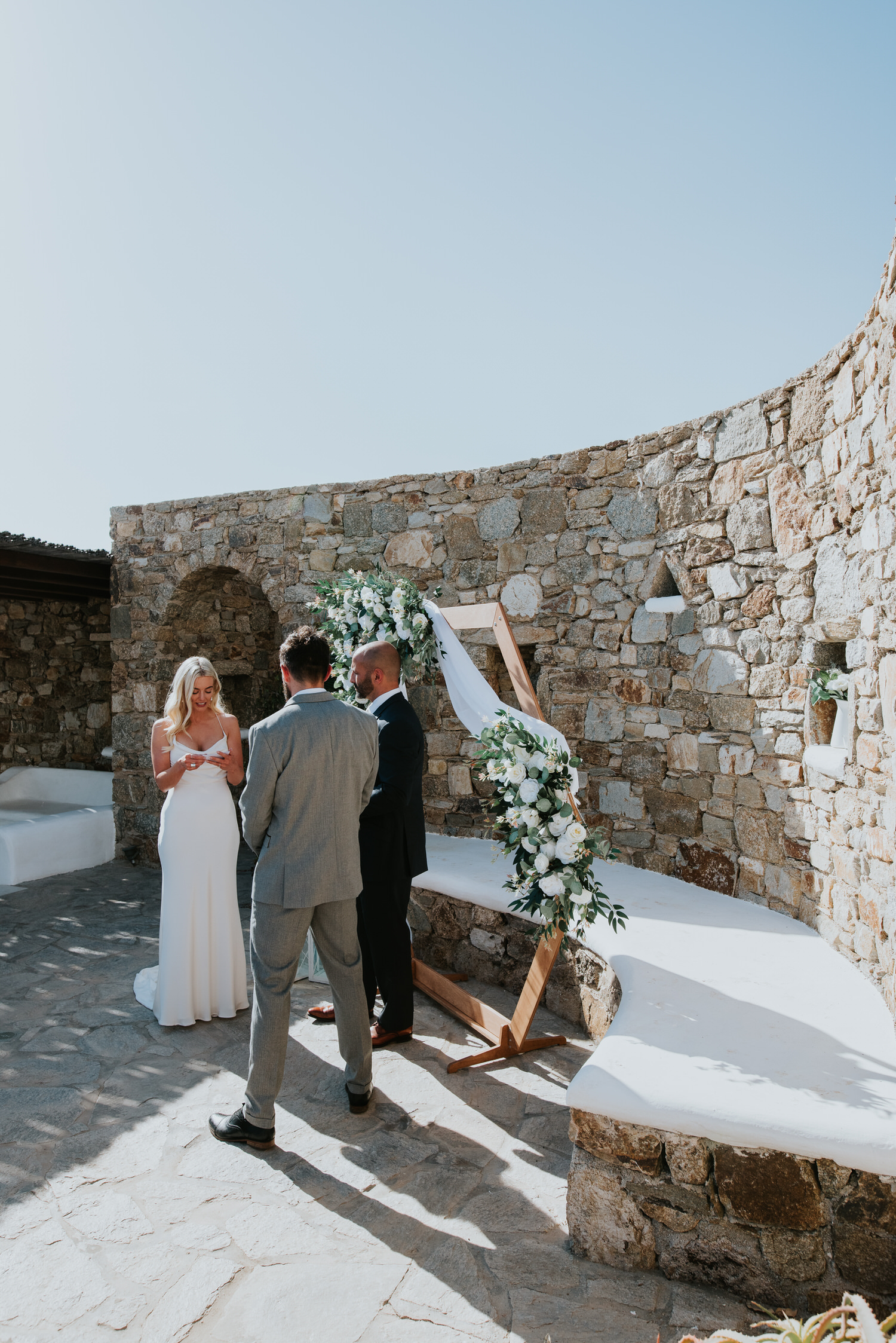 Mykonos wedding photographer: bride reading her vows as groom and celebrant watch her in front of the floral arch and the rock wall.