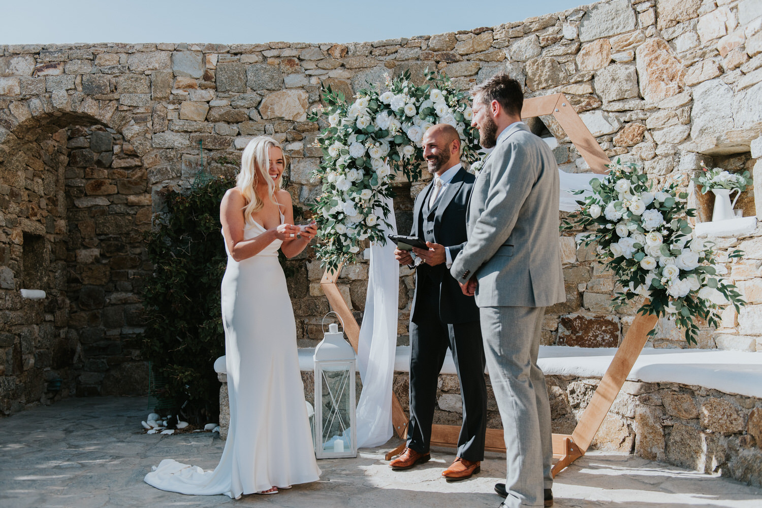 Mykonos wedding photographer: bride laughing holding her vows in front of the floral arch as celebrant and groom watch her.