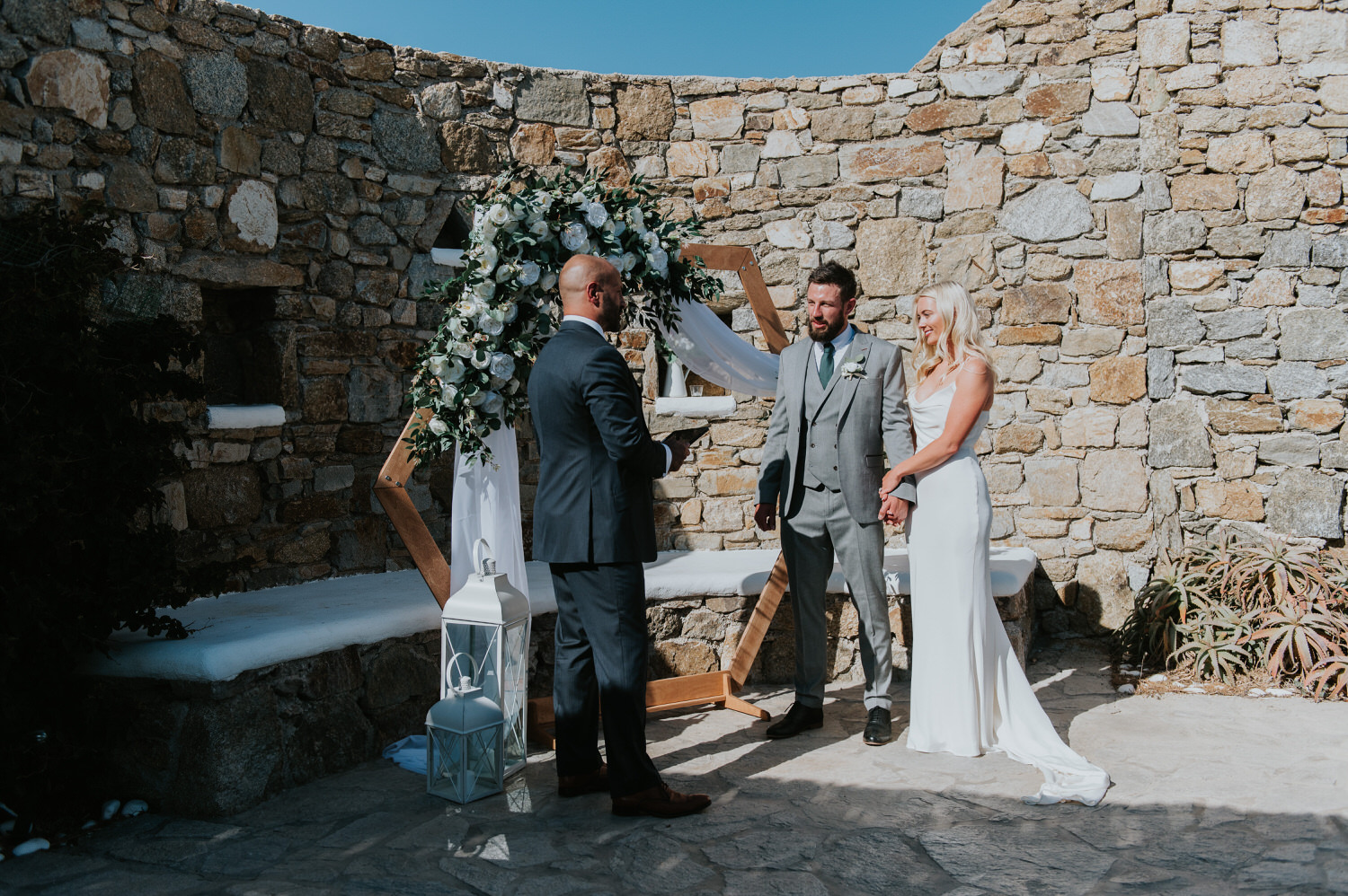 Mykonos wedding photographer: bride and groom holding hands in front of floral arch looking at the celebrant with the rock wall in the background.