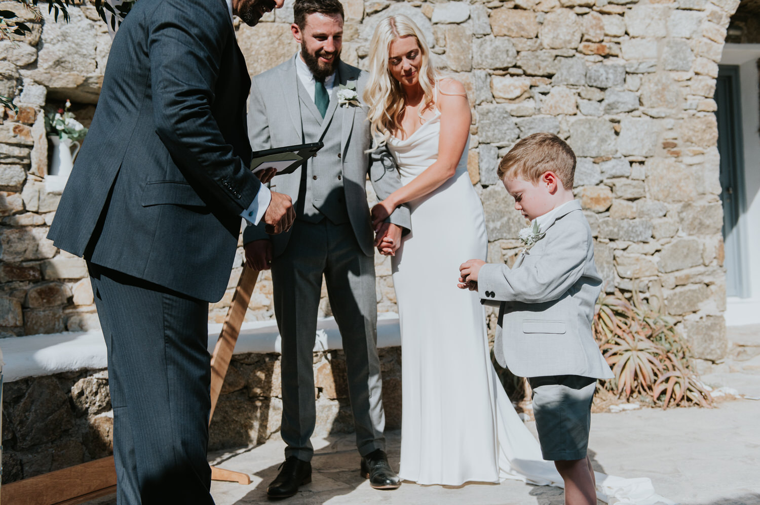 Mykonos wedding photographer: celebrant taking rings from the little ring bearer as bride and groom smile looking at him.
