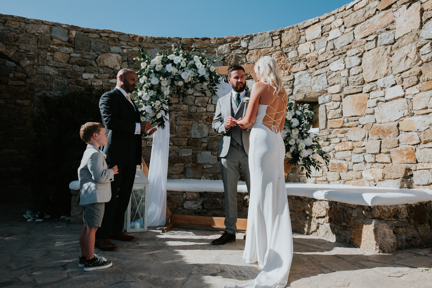 Mykonos wedding photographer: celebrant, ring bearer, bride and groom in front of the floral arch exchanging the rings in afternoon sun.