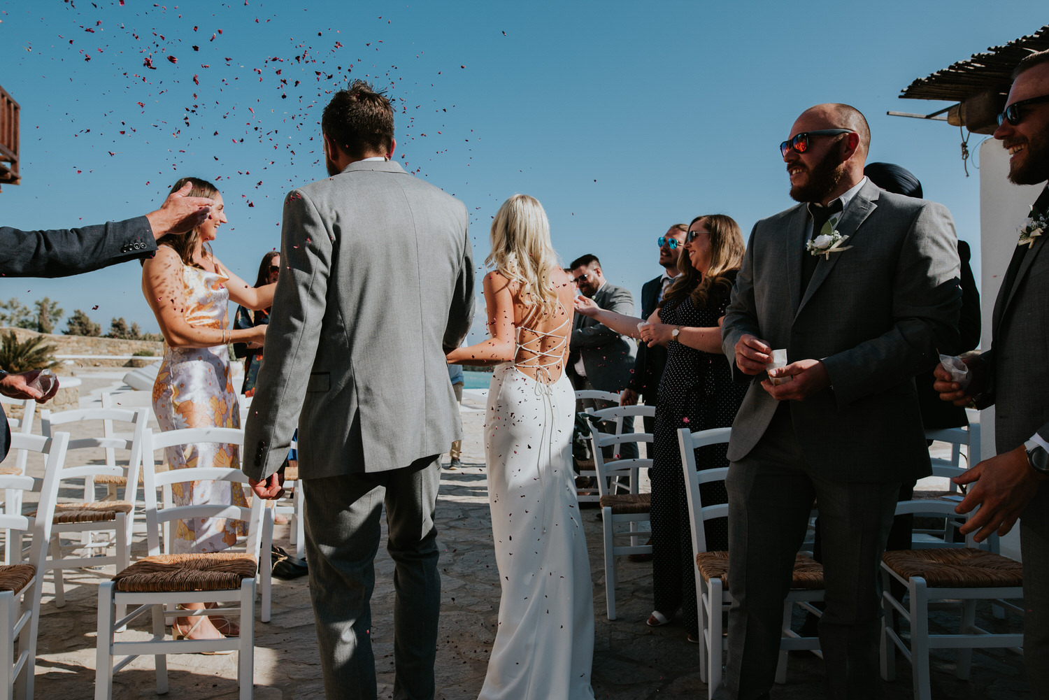 Mykonos wedding photographer: bride and groom shot from behind exiting the aisle holding hands showered in dry petals.