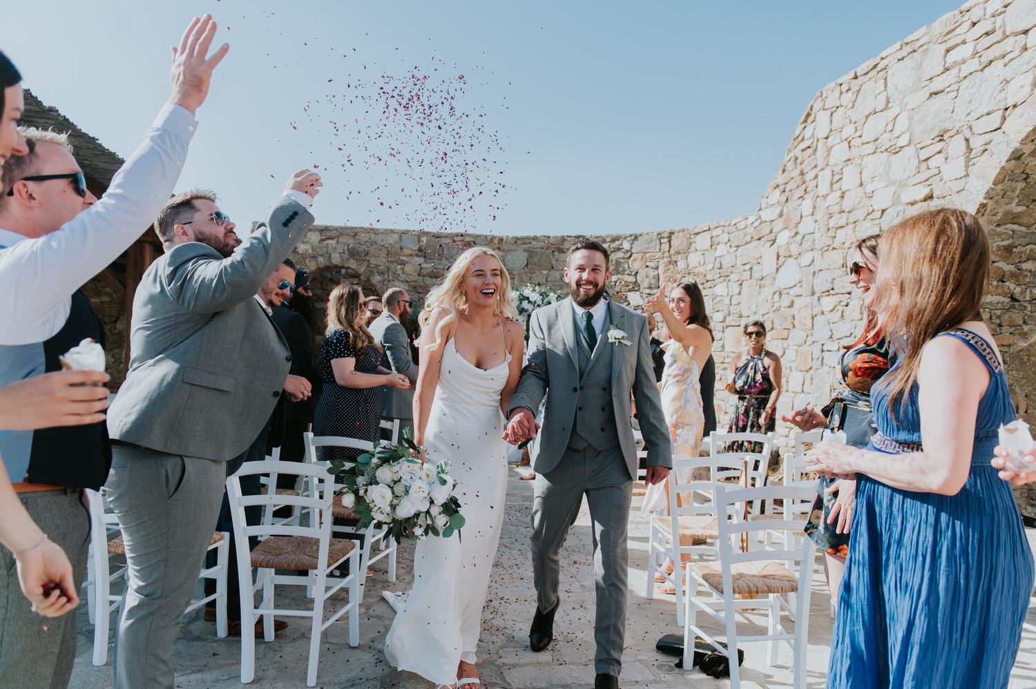 Mykonos wedding photographer: friends laughing and showering bride and groom on their exit from the aisle holding hands and laughing.