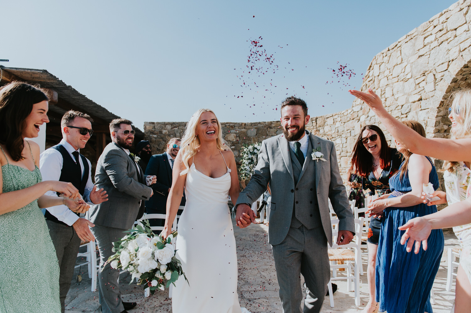 Mykonos wedding photographer: bride and groom exiting the aisle holding hands and laughing showered in dry petals by friends around them.