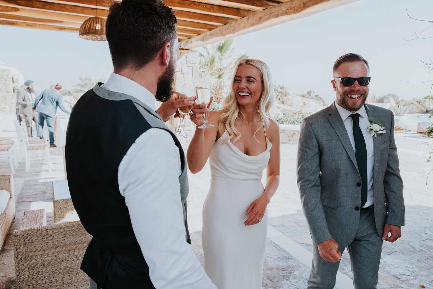 Mykonos wedding photographer: bride and groom smiling clinking champagne glasses with one of the guests.