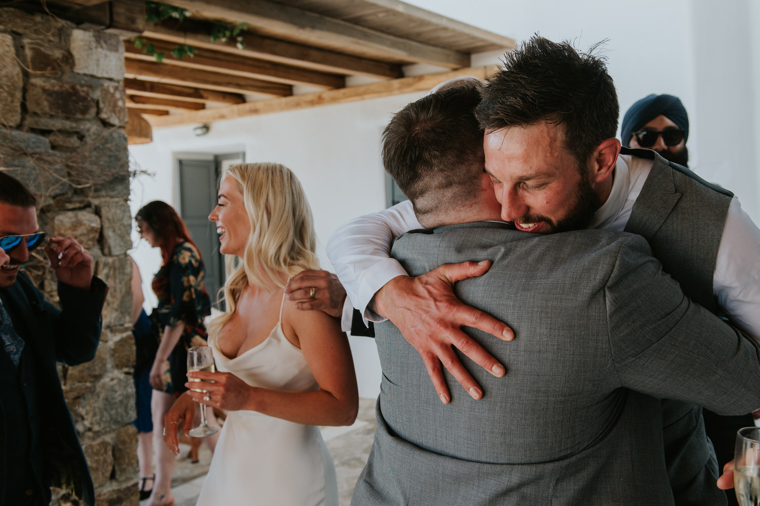Mykonos wedding photographer: groom hugging his best man with bride next to him and a glass of champagne in her hand.