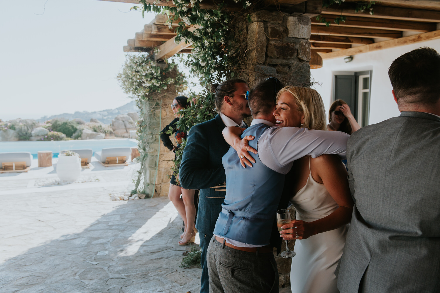 Mykonos wedding photographer: bride hugging a guest smiling with a glass of champagne in her hand.