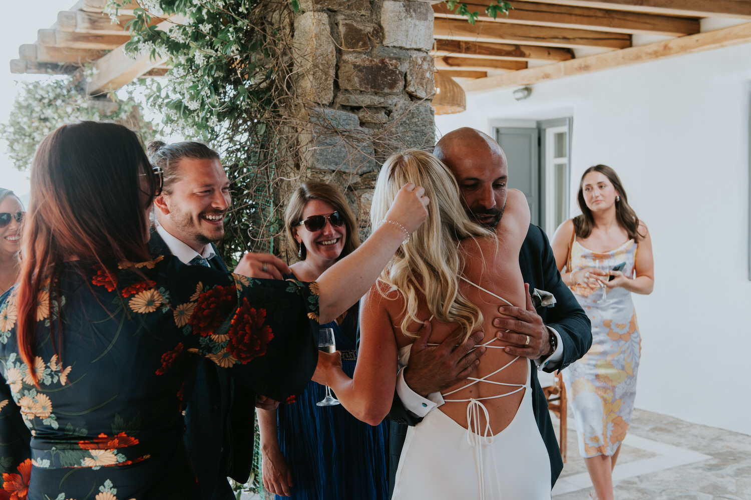 Mykonos wedding photographer: bride with her lace up back of the dress hugging a guest surrounded by other smiling faces.