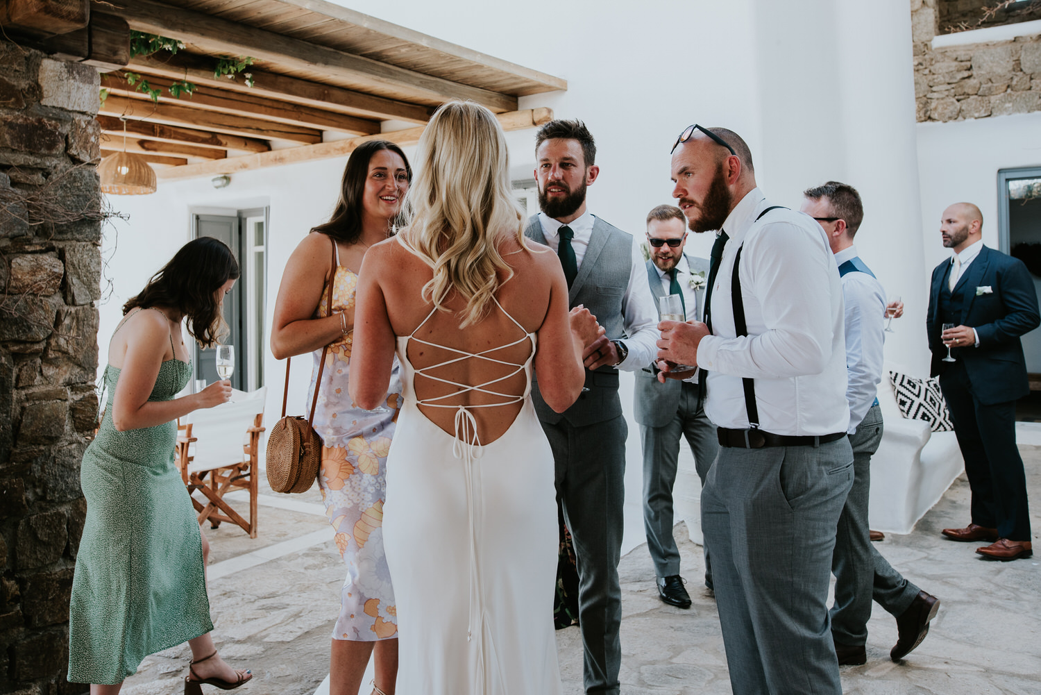Mykonos wedding photographer: bride with her back towards the camera talking to the groom and friends in front of the villa.