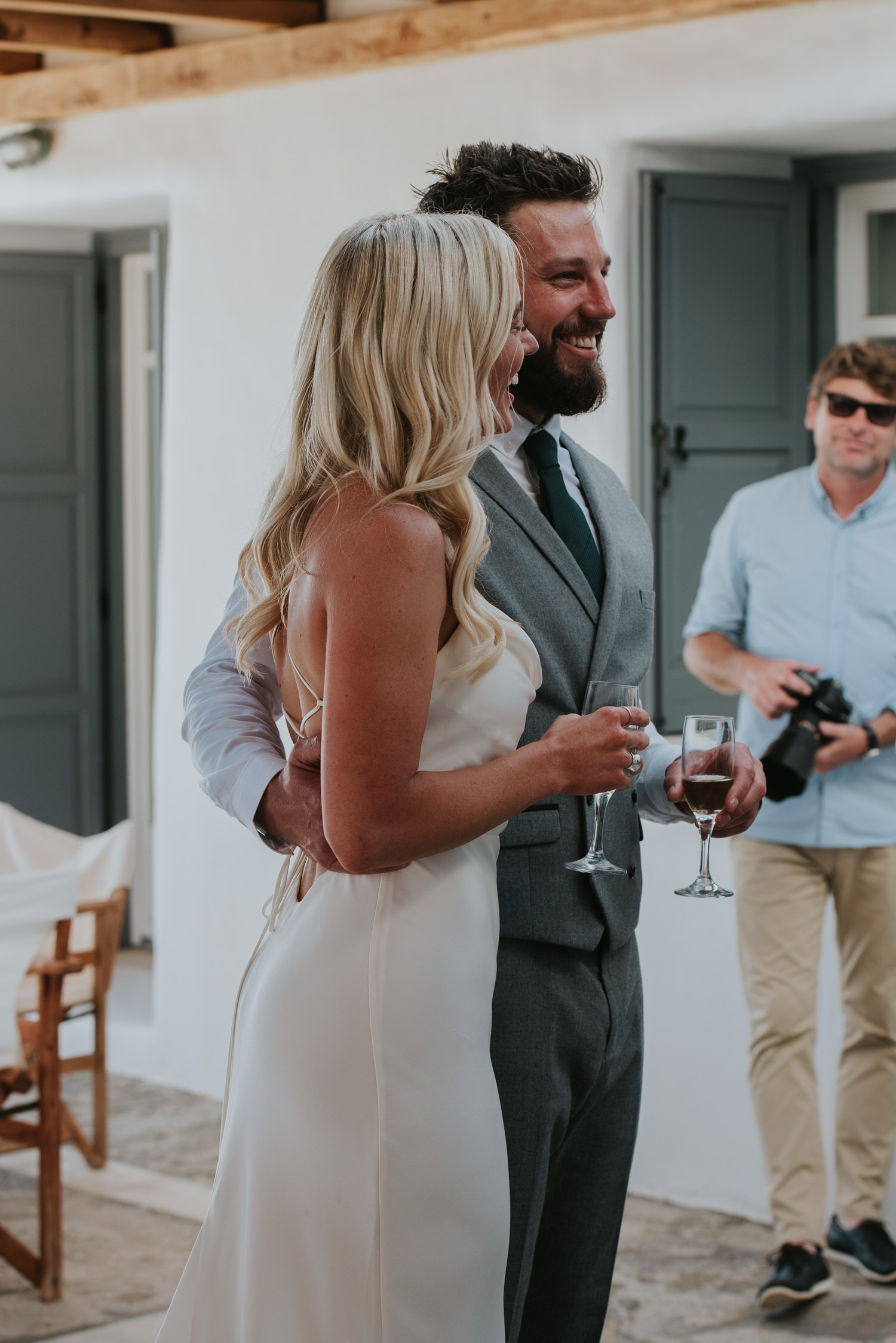 Bride and groom laughing holding champagne glasses in the shade with Mykonos wedding photographer smiling in the background.