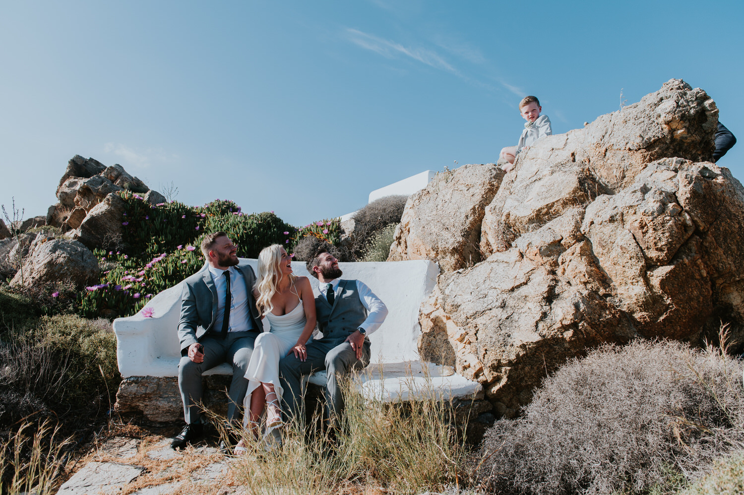Mykonos wedding photographer: bride, groom and best man sat on a bench nestled in the rocks looking up towards the rock where a kid is sat.