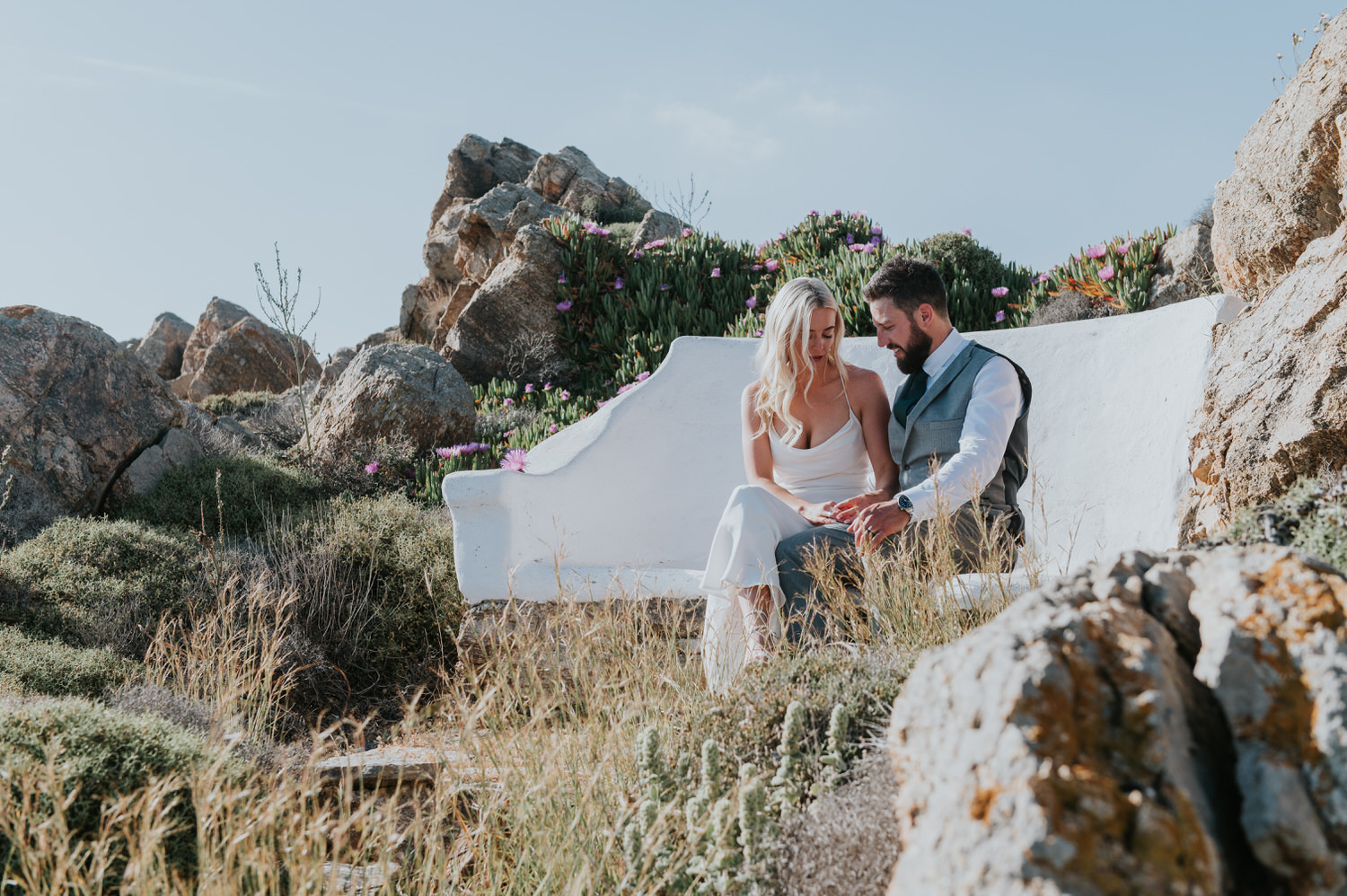 Mykonos wedding photographer: bride and groom sat on a bench nestled in the rocks looking at their hands in the afternoon light.