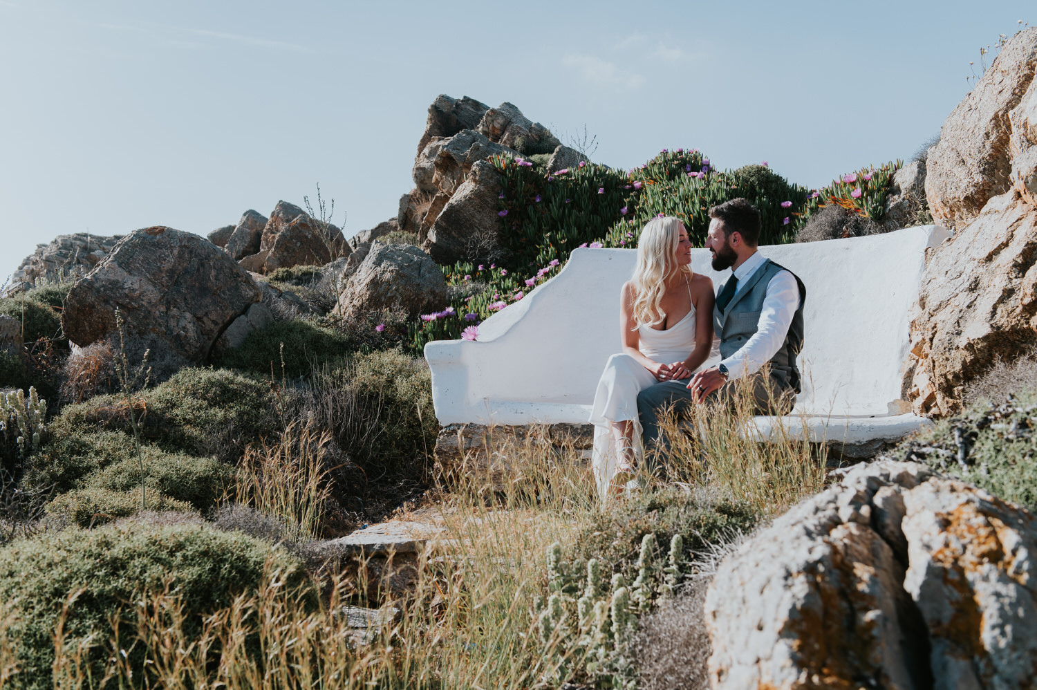 Mykonos wedding photographer: bride and groom sat on a bench nestled in the rocks looking at each other in the afternoon light.