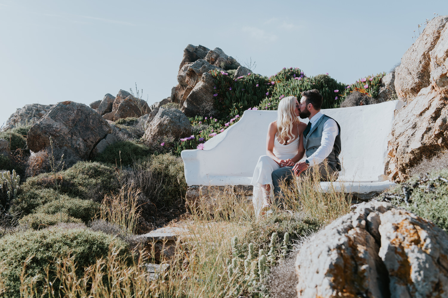 Mykonos wedding photographer: bride and groom sat on a bench nestled in the rocks kissing in the afternoon light.