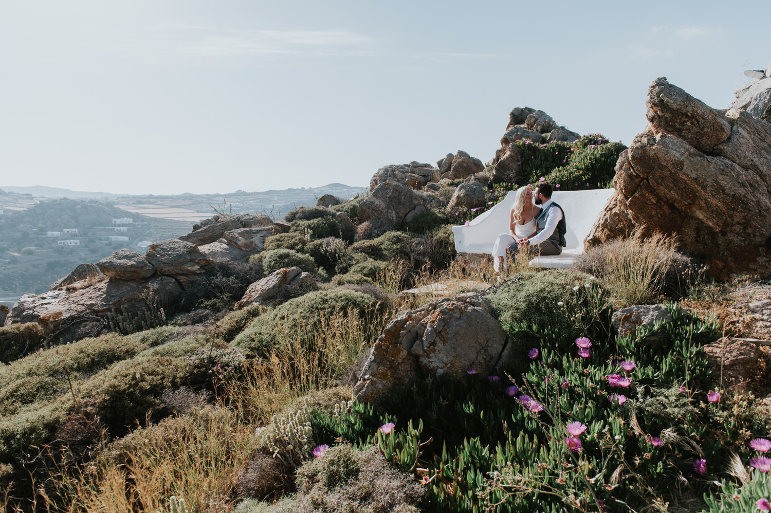 Mykonos wedding photographer: bride and groom sat on a bench nestled in the rocks surrounded by the landscape in the afternoon light.