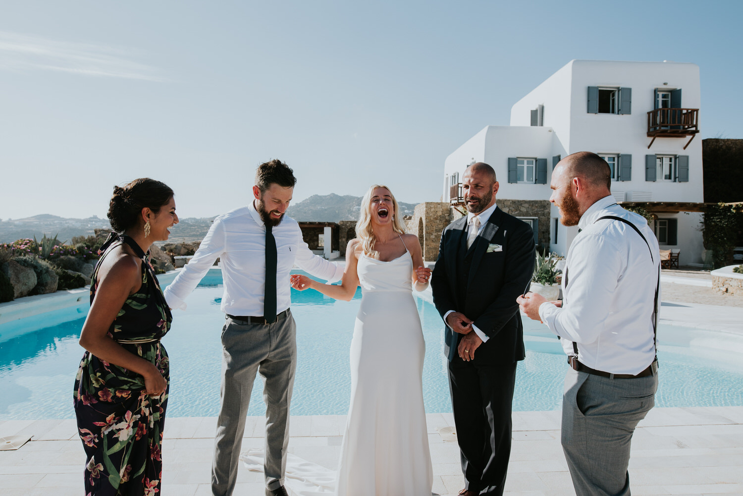 Mykonos wedding photographer: bride and groom with their friends laughing next to the pool in afternoon light.