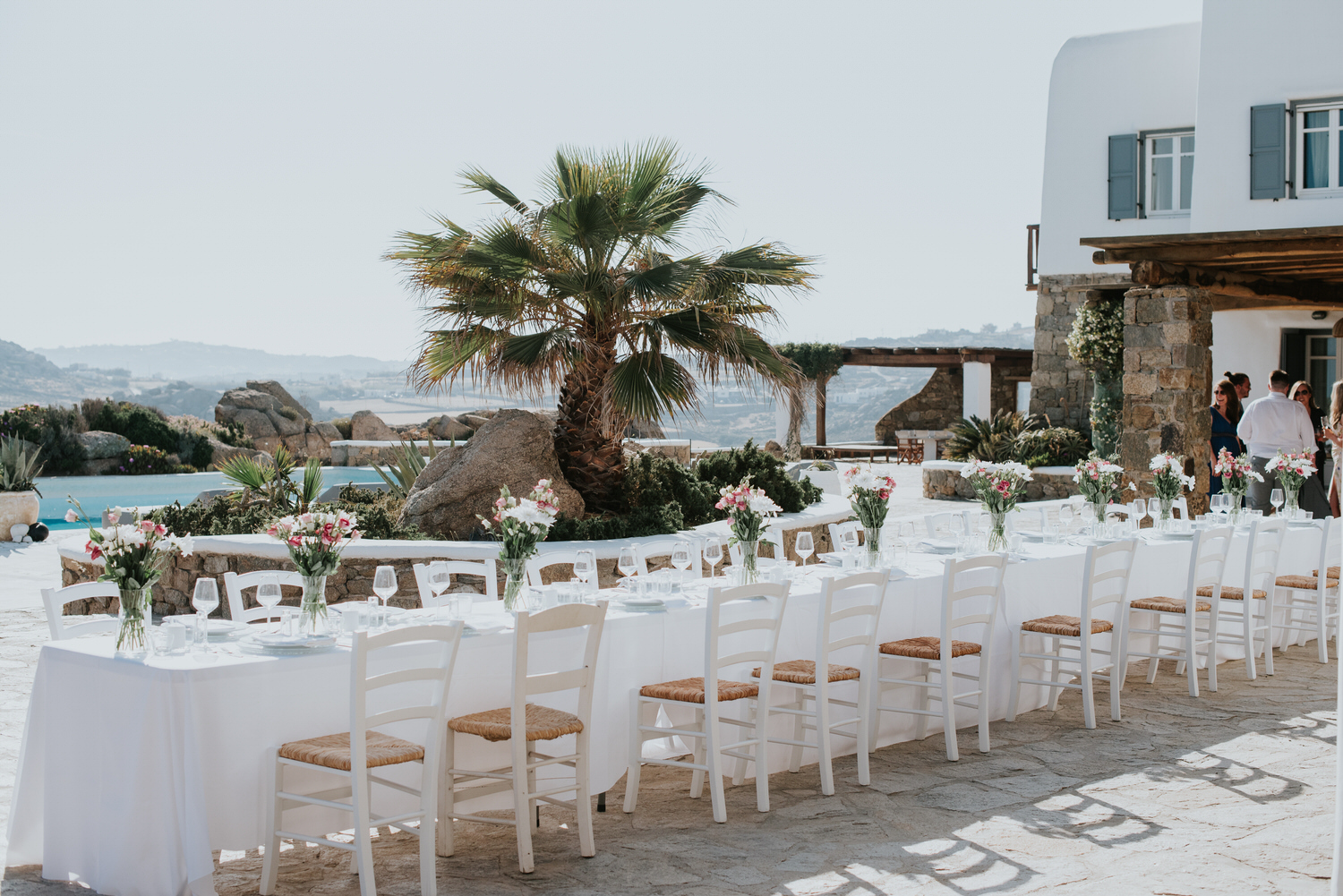 Mykonos wedding photographer: panoramic photo of long reception table setup in the afternoon light with flower arrangements, villa and guests in the background.