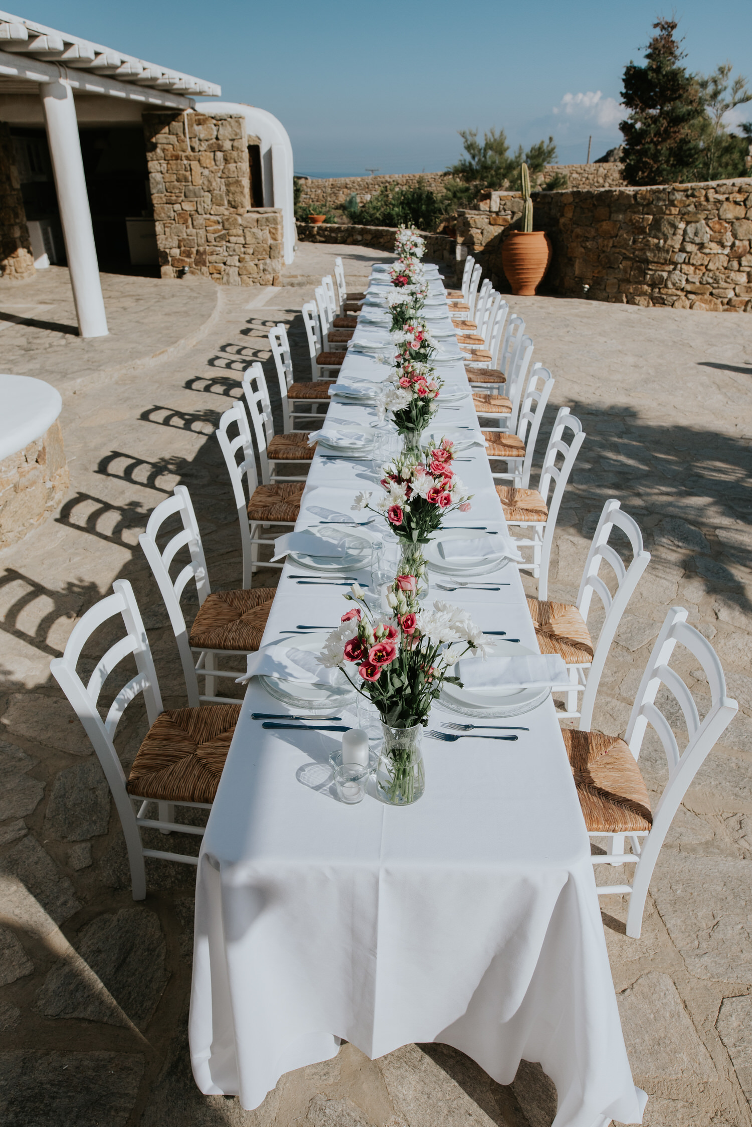 Mykonos wedding photographer: long reception table setup in the afternoon light with flower arrangements and guests and the villa in the background.