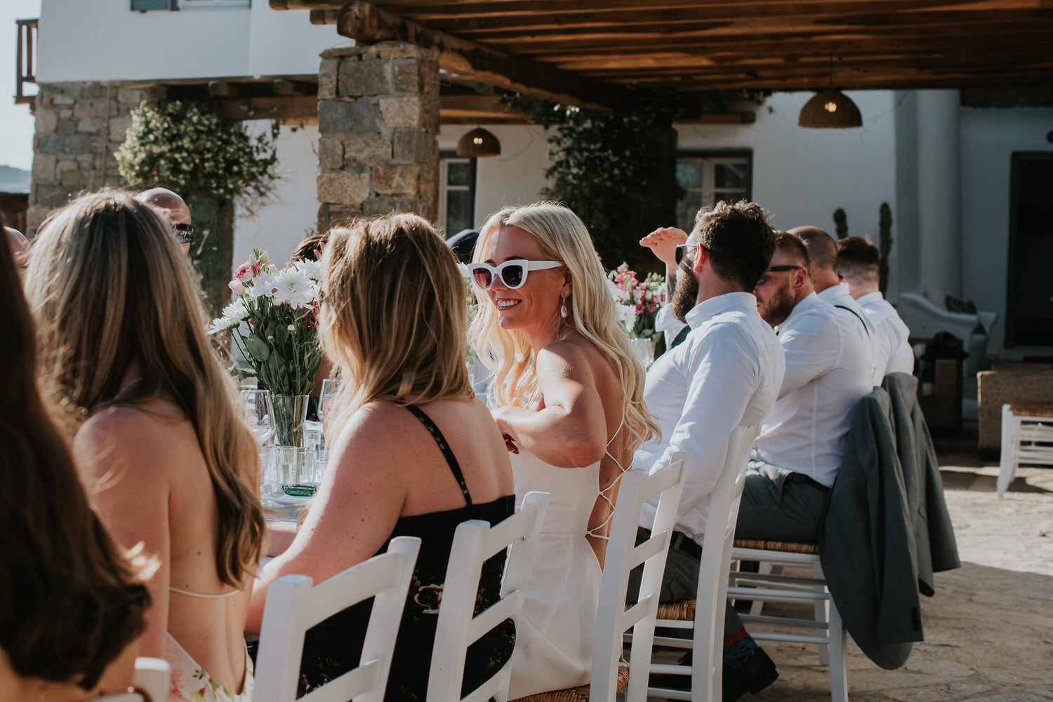 Mykonos wedding photographer: accent on bride at the long reception table surrounded by guests in the afternoon sun.